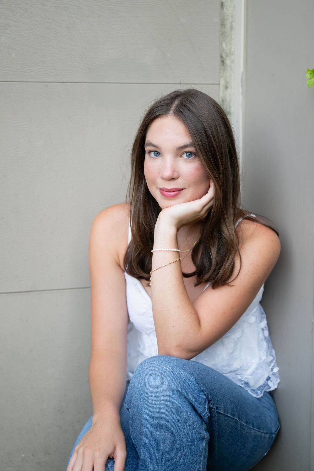 Young woman with brown hair in white tank top and jeans sits against gray wall, resting chin on hand.