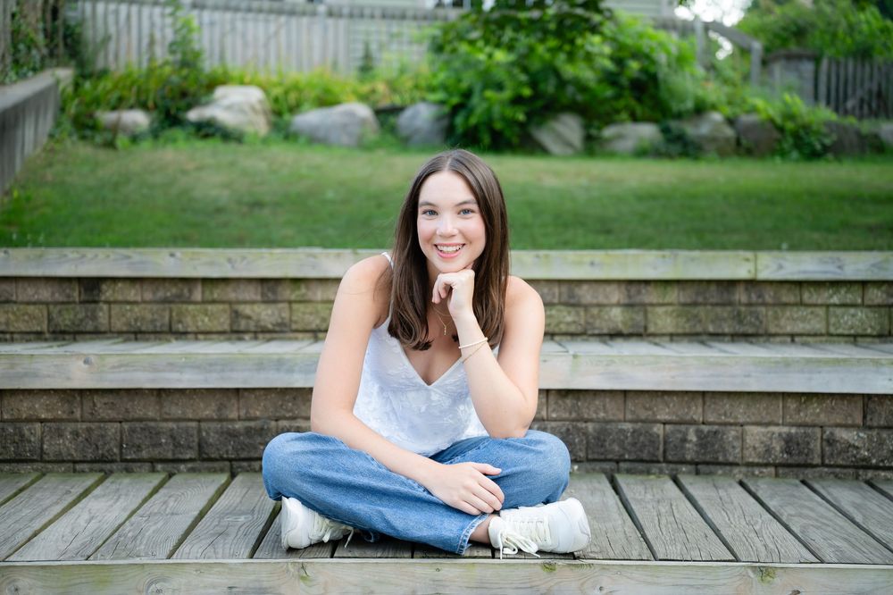 Young woman smiling, sitting cross-legged on wooden steps outdoors, wearing jeans and white top, with green garden background.