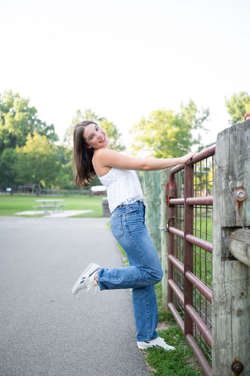 Young woman in jeans and white top poses playfully by a rustic metal gate in a scenic park setting.
