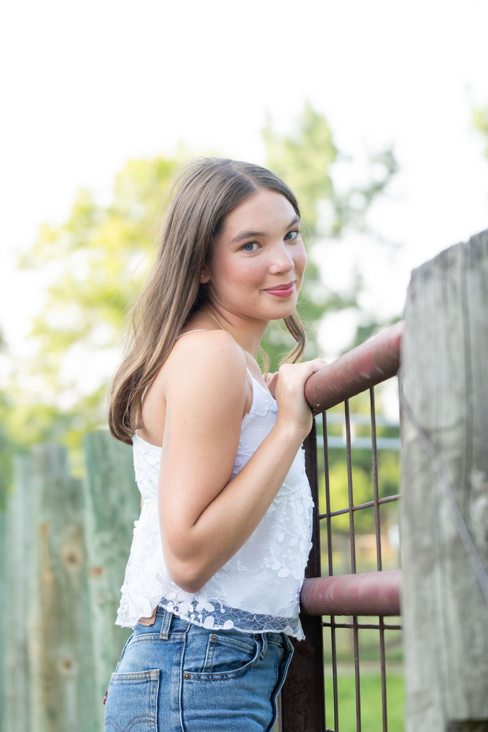 Young woman in white lace top and jeans leaning on rustic metal fence outdoors with green trees in background.