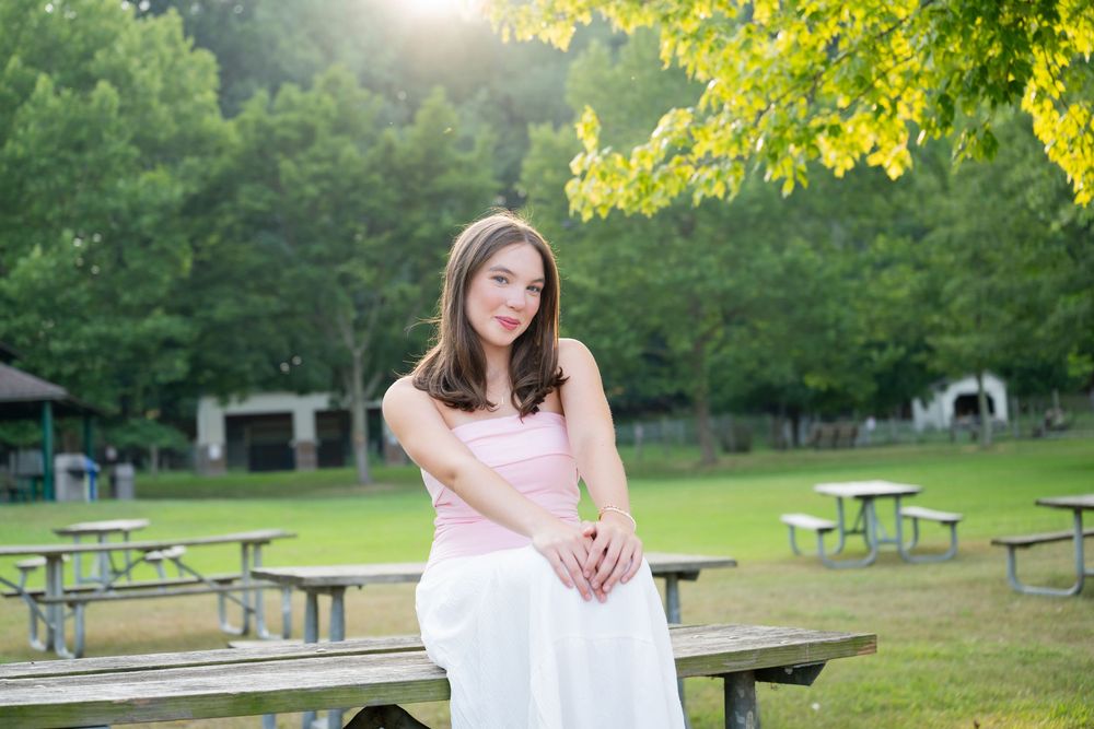 Young woman in pink top and white skirt sitting on picnic table in lush green park setting.