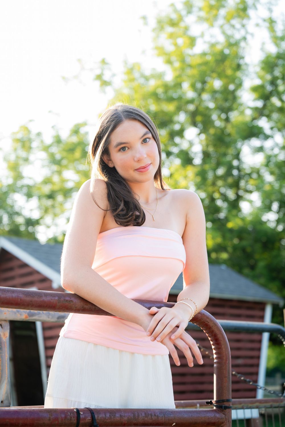 Young woman in pink tube top and white skirt leans on rustic railing outdoors with green trees behind her.