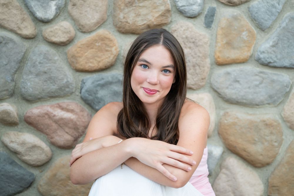 Young brunette woman smiling, arms crossed, sitting against a rustic stone wall in a pink top.