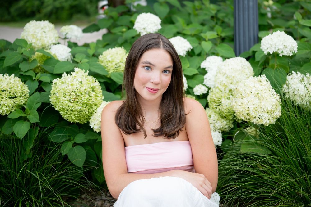 Young brunette woman in pink strapless dress sitting in front of lush green hydrangea bushes with white blooms.