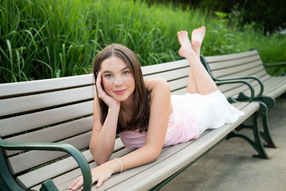 Young woman in pink dress lying on a wooden park bench with lush green grass in the background.