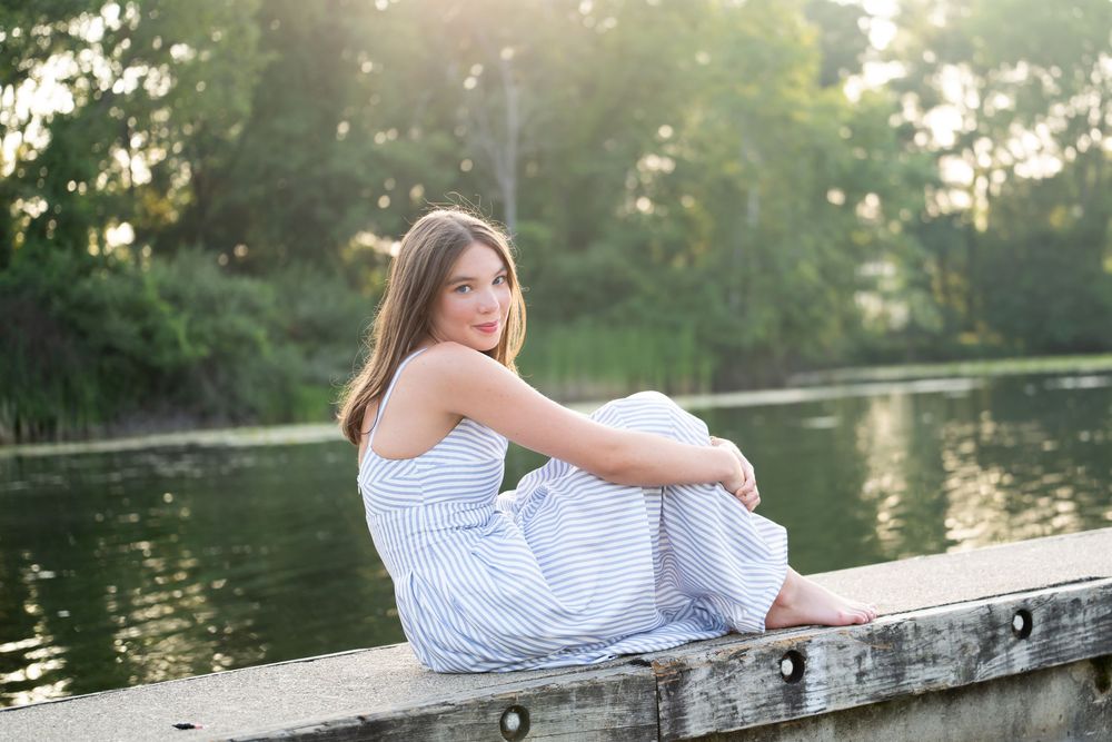 Young woman in striped dress sitting on dock by serene lake surrounded by lush green trees in golden sunlight.