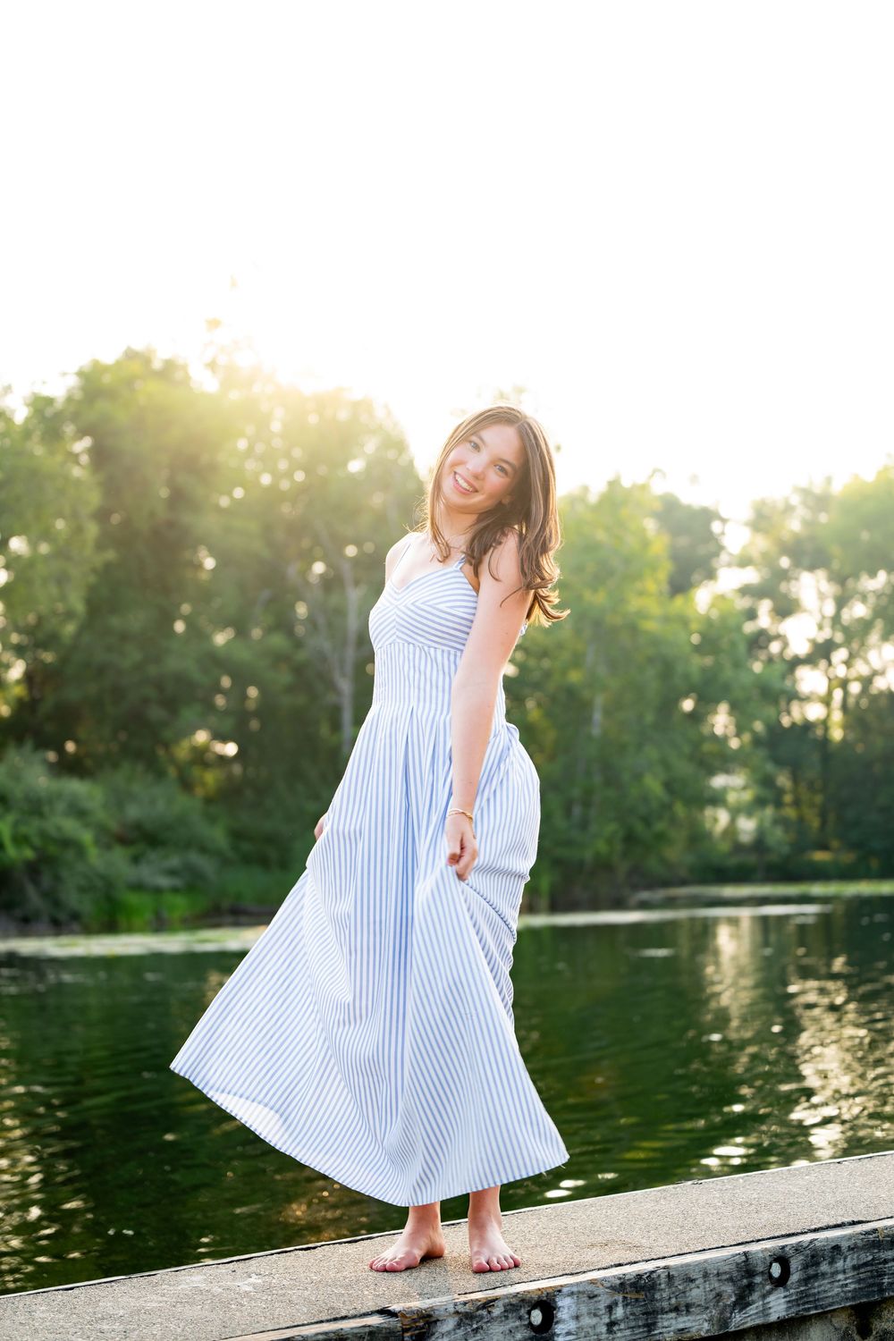 Young woman in flowing blue striped maxi dress standing barefoot on dock by sunlit lake surrounded by green trees.