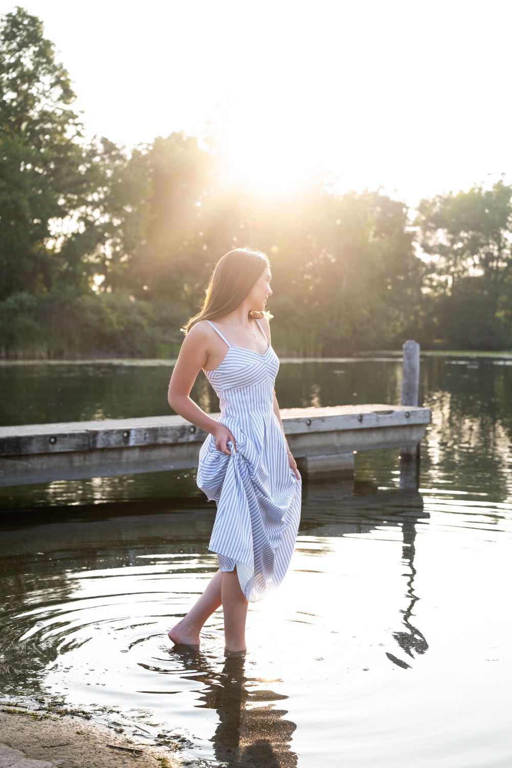 Young woman in blue striped sundress wading in calm lake at golden sunset near wooden dock.