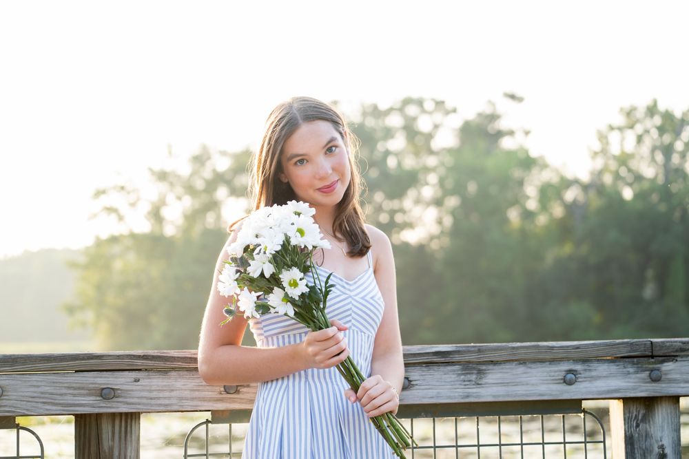 Young woman in striped dress holding white daisy bouquet outdoors in soft sunlight near wooden fence.
