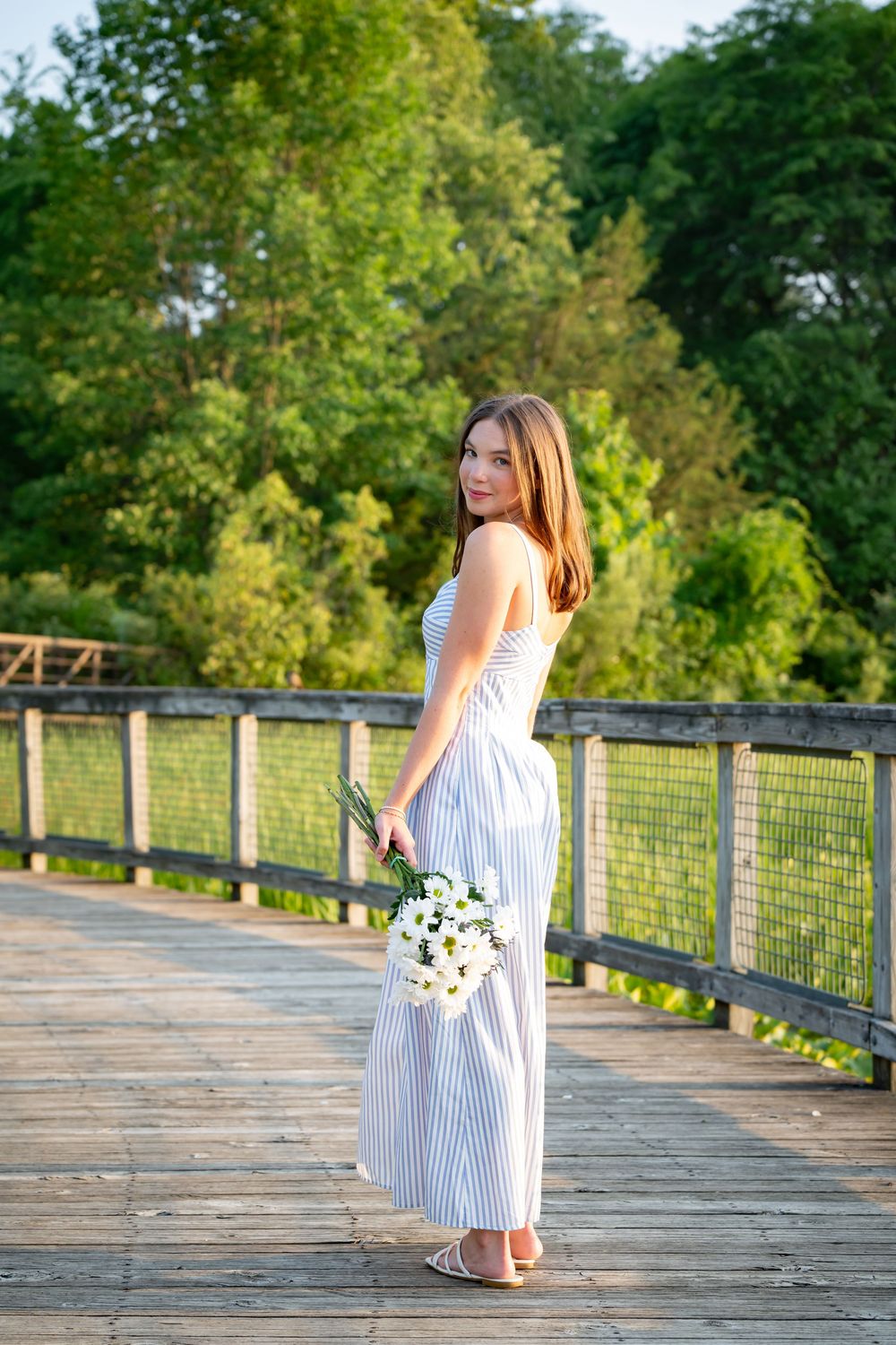 Young woman in striped maxi dress holding white flowers on a wooden bridge surrounded by lush green trees.