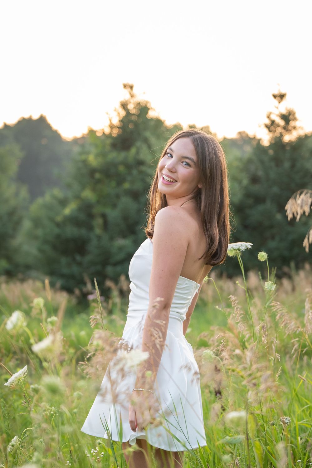 Young woman in white strapless dress smiling in a sunlit meadow with wildflowers and green trees at golden hour.