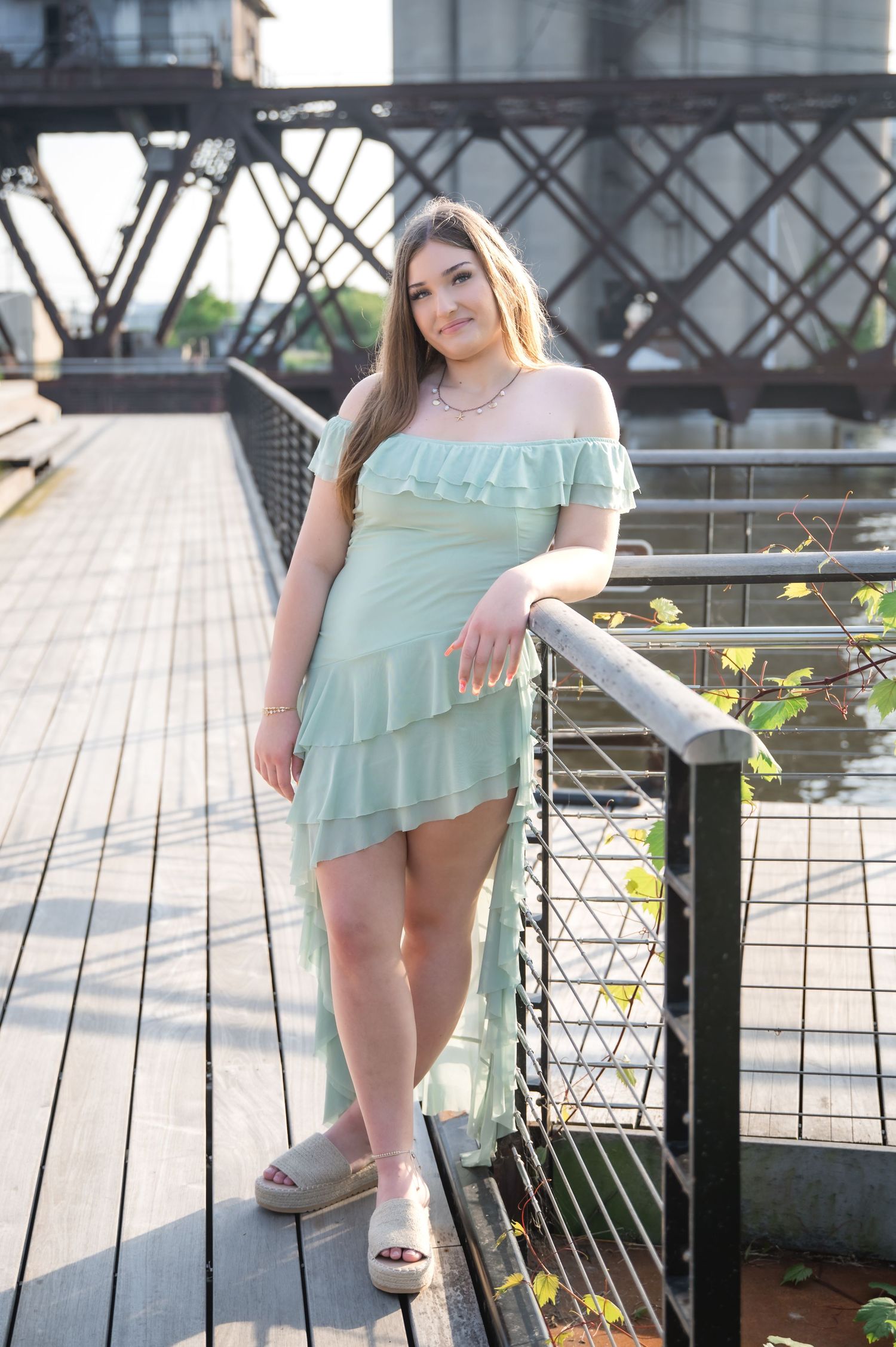 Young woman in mint green off-shoulder ruffle dress posing on a wooden bridge with metal railings outdoors.