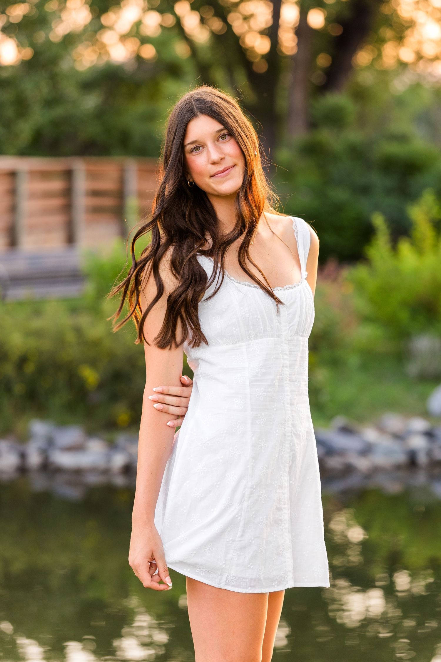 Young woman with long dark hair in white sundress poses near a pond in a lush garden at golden hour.