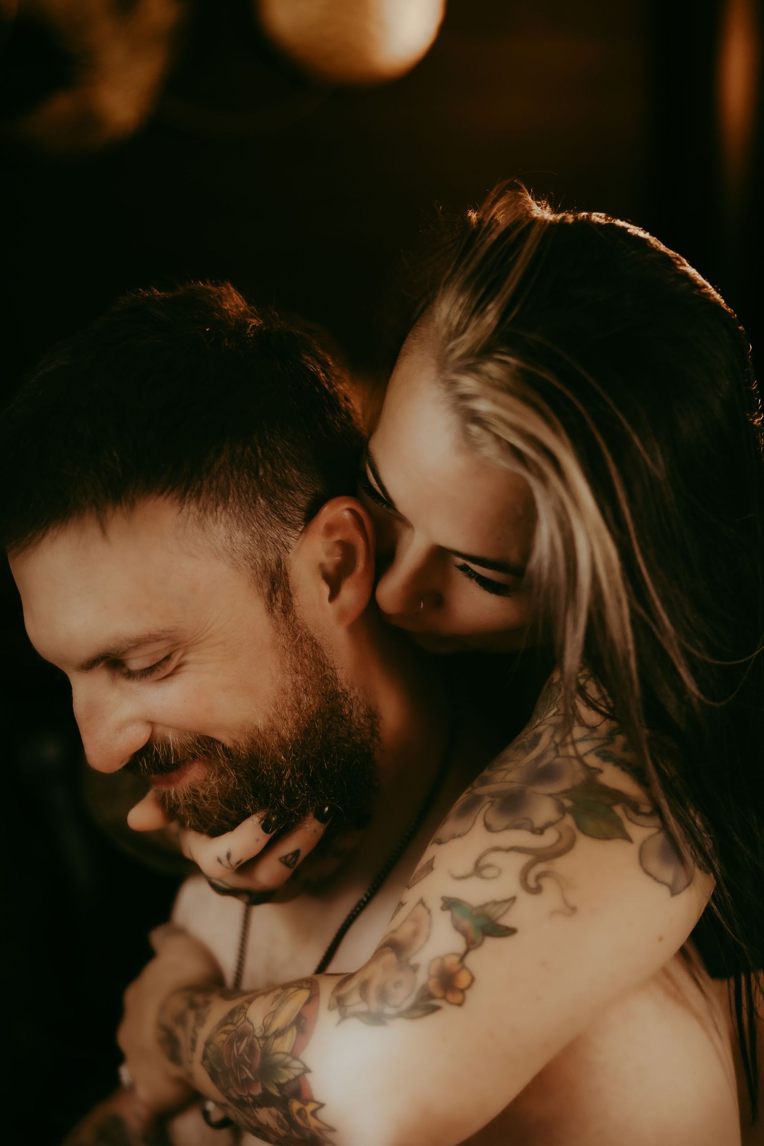 A smiling bearded tattooed man embraced from behind by a tattooed blonde woman in a warm, dimly lit setting.