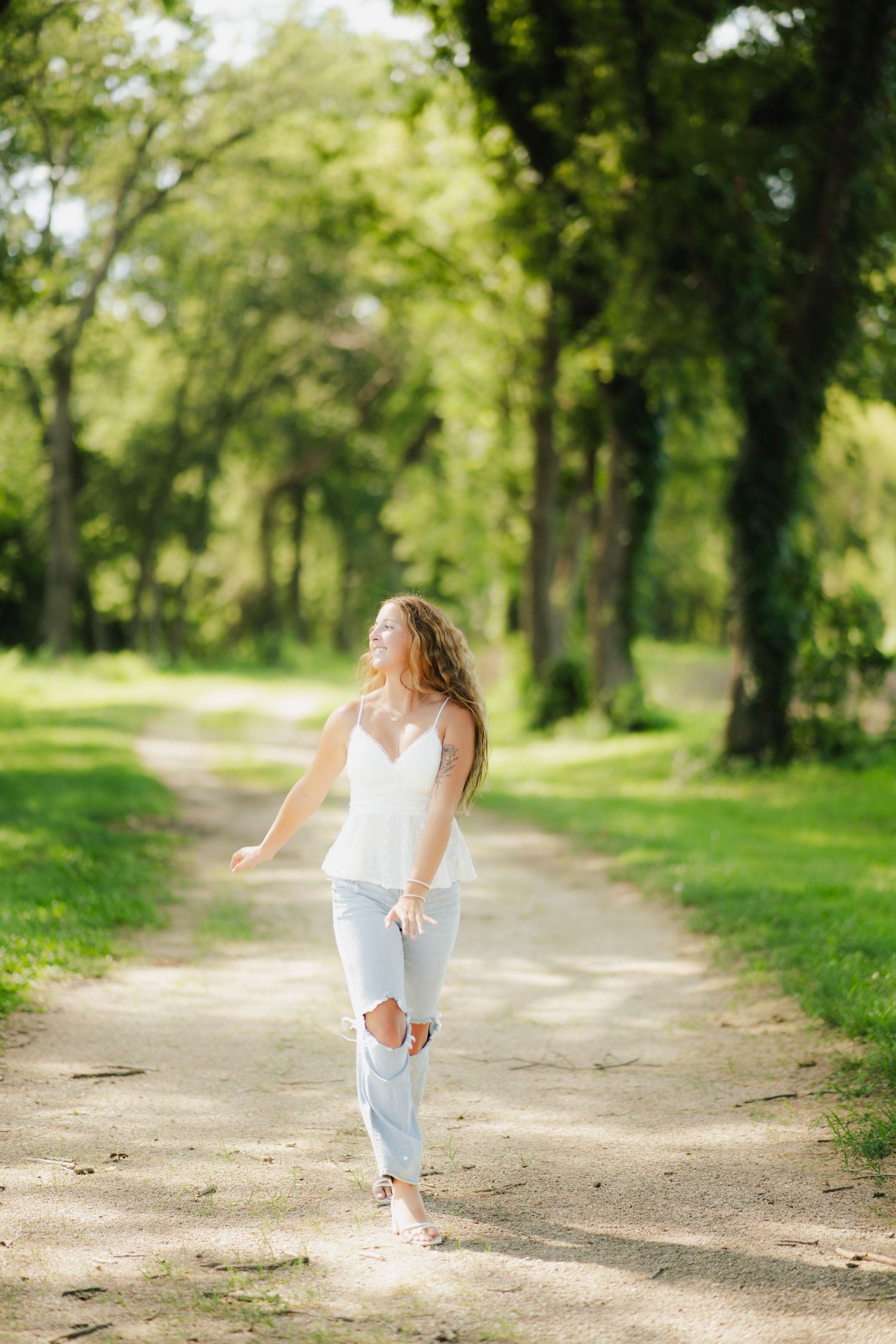 Young woman in white top and ripped jeans walking on a sunny park path lined with green trees.