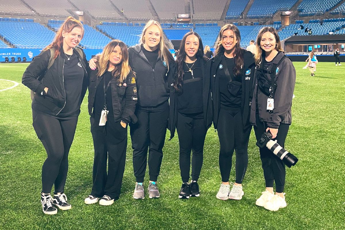 Six women in black outfits pose together on a stadium field with blue seats visible in the background.