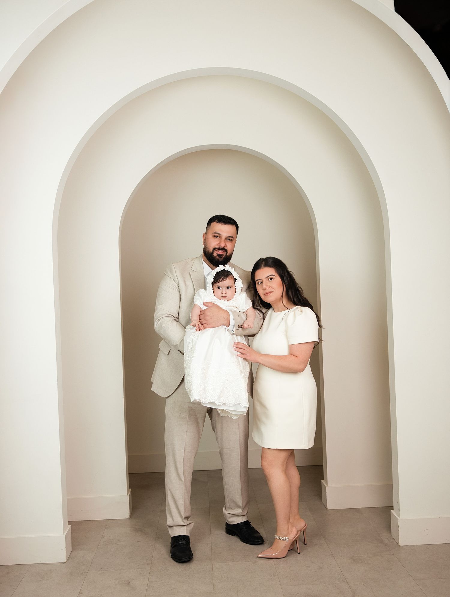 Family portrait with parents in white and beige holding baby in christening gown under elegant arched backdrop.