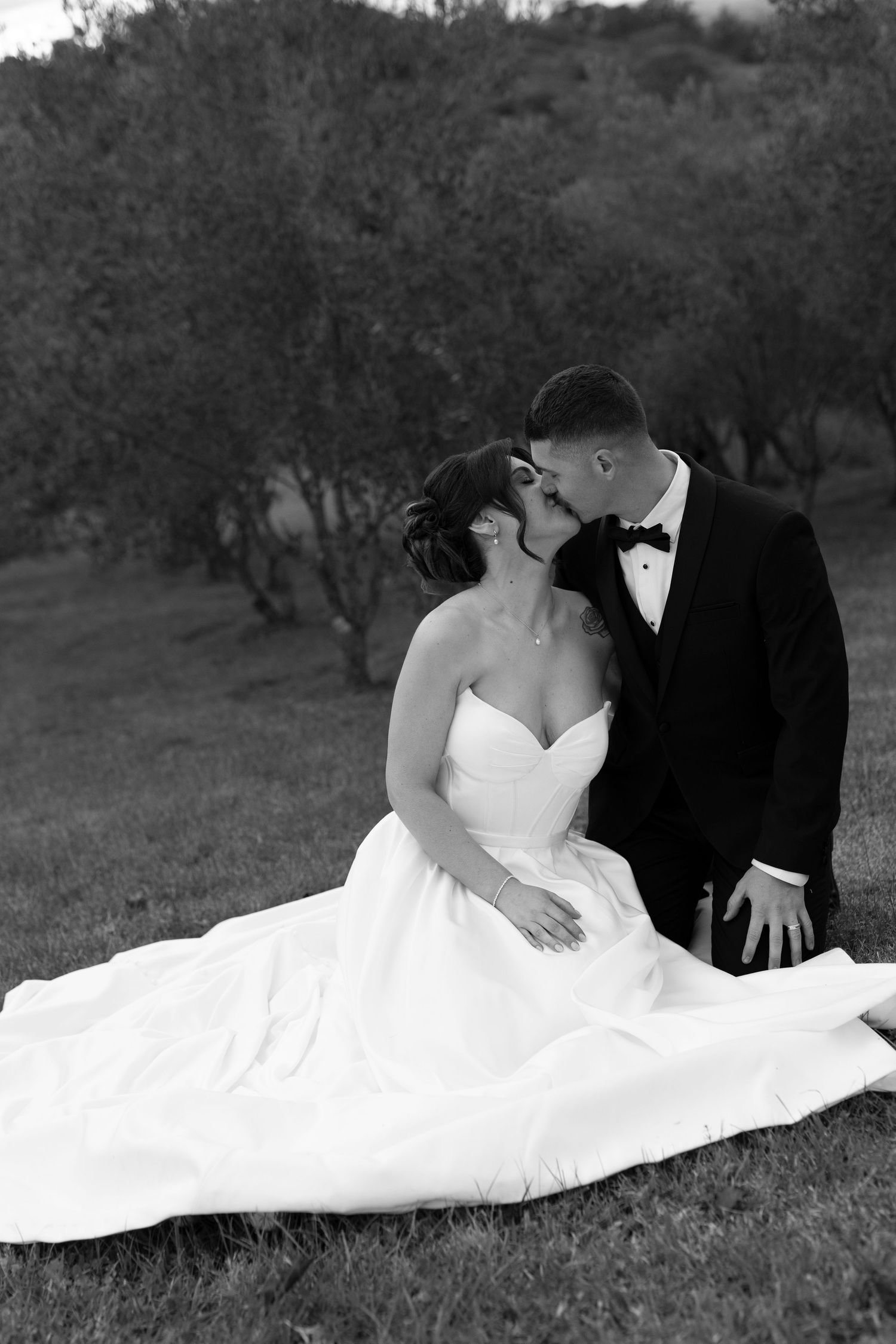 Bride in white gown sits beside groom in black tuxedo lying on grass, with olive trees in background.