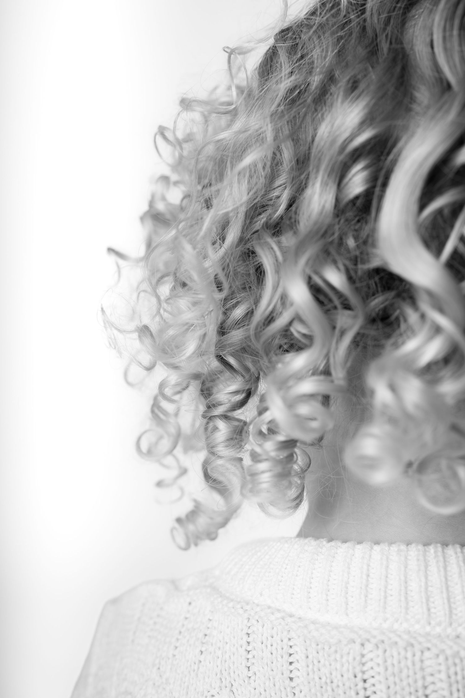 Black and white photo of a woman with voluminous curly blonde hair wearing a knit sweater, viewed from behind.