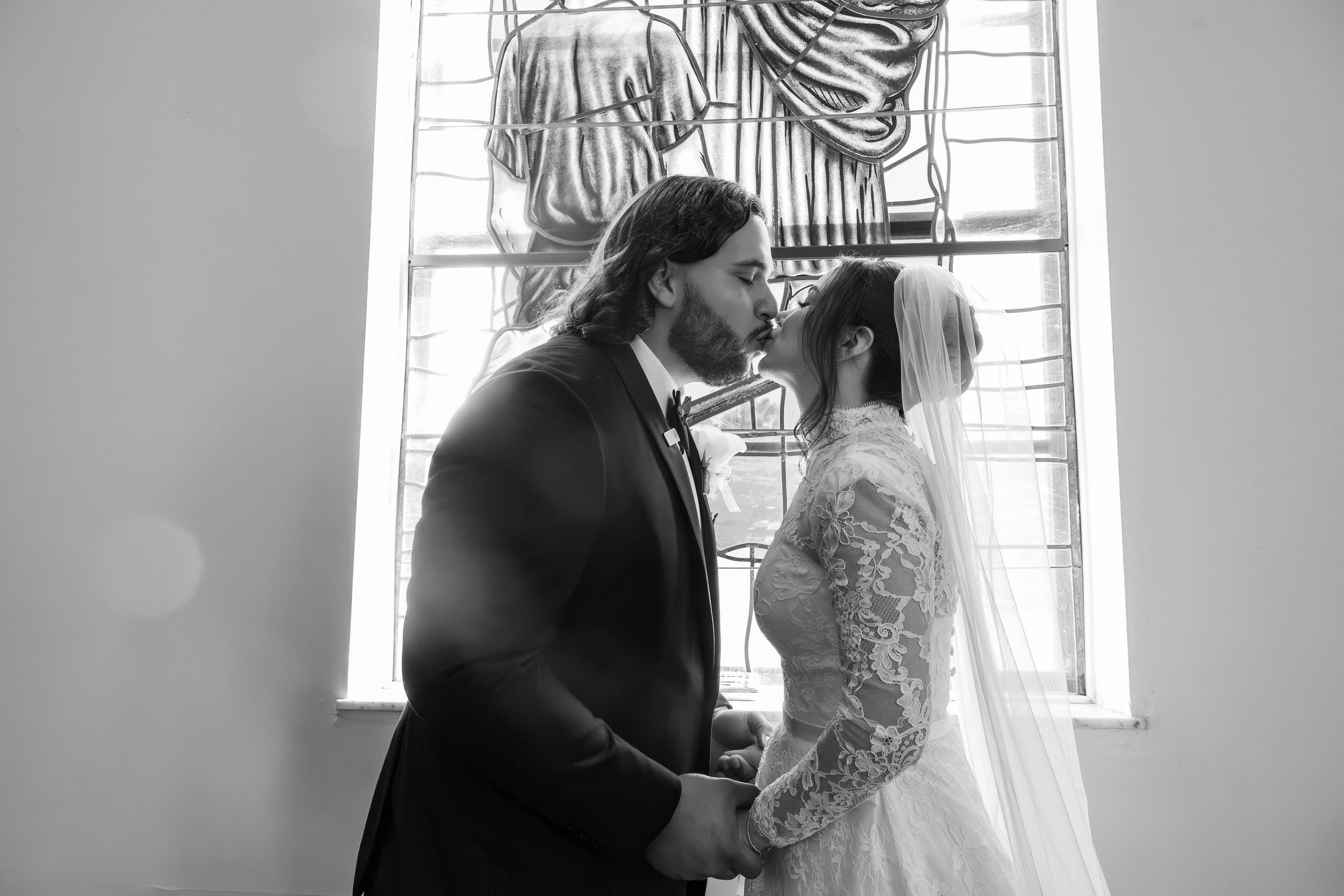 Black and white photo of a bride and groom holding hands and sharing a romantic moment in front of a stained glass church window.