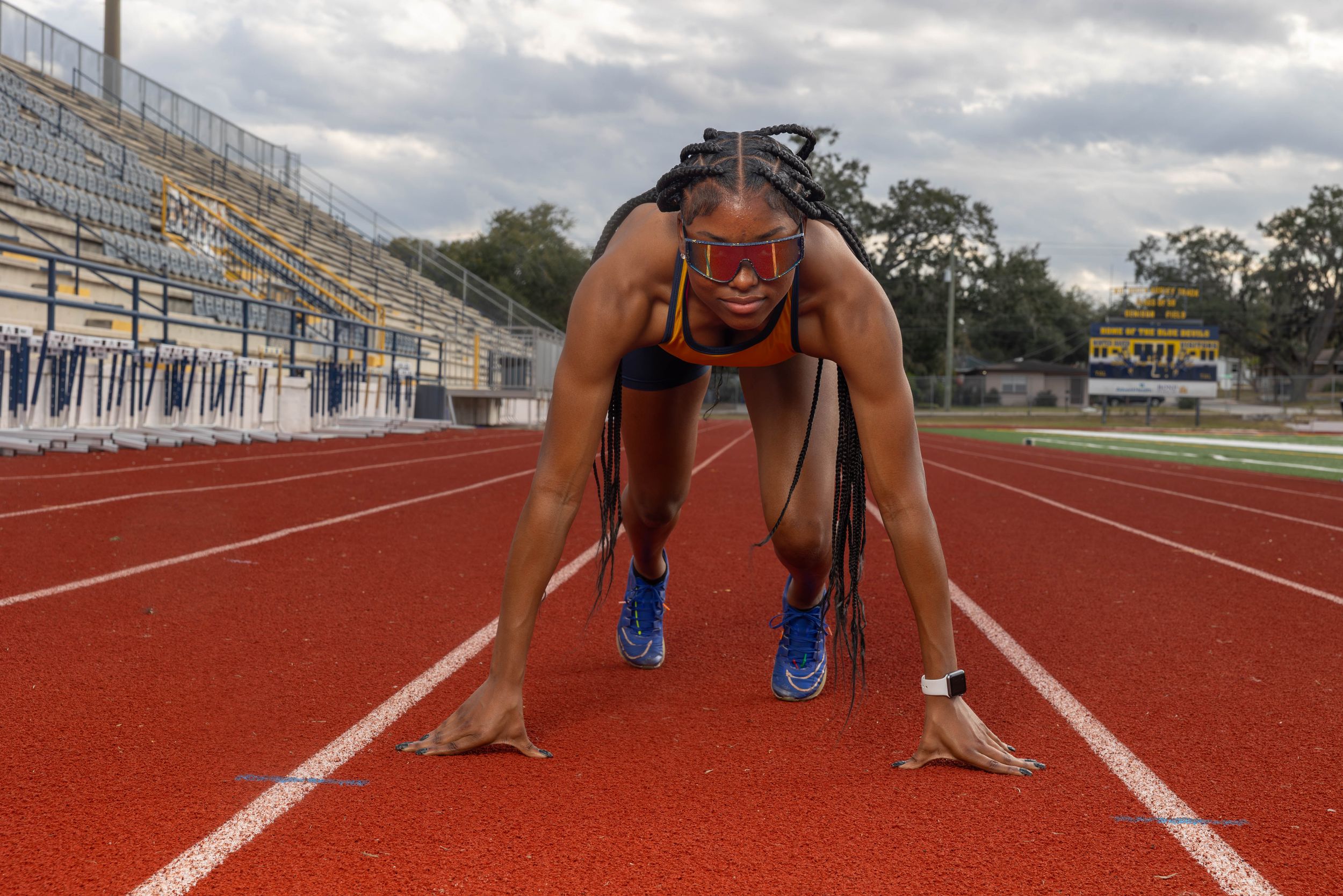 Female sprinter in starting position on red track, wearing goggles and athletic gear, stadium bleachers in background.
