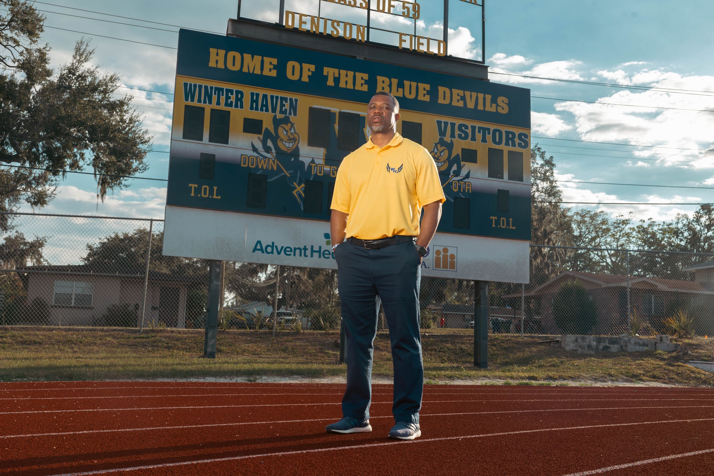 Man in yellow shirt stands on track at Winter Haven Blue Devils football stadium with scoreboard visible.