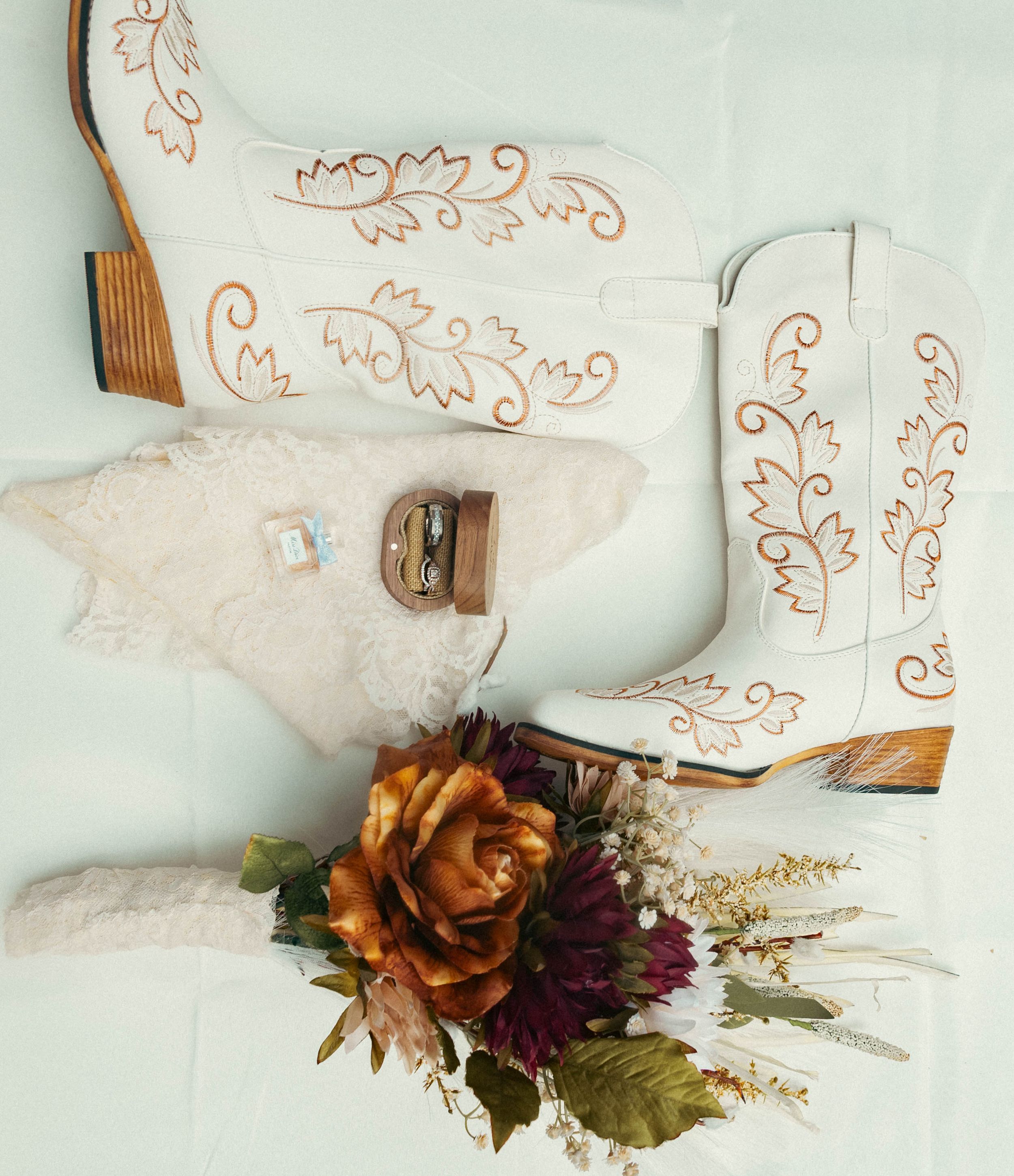 White cowboy boots with gold embroidery displayed with wedding rings and a fall floral bouquet featuring burnt orange roses.
