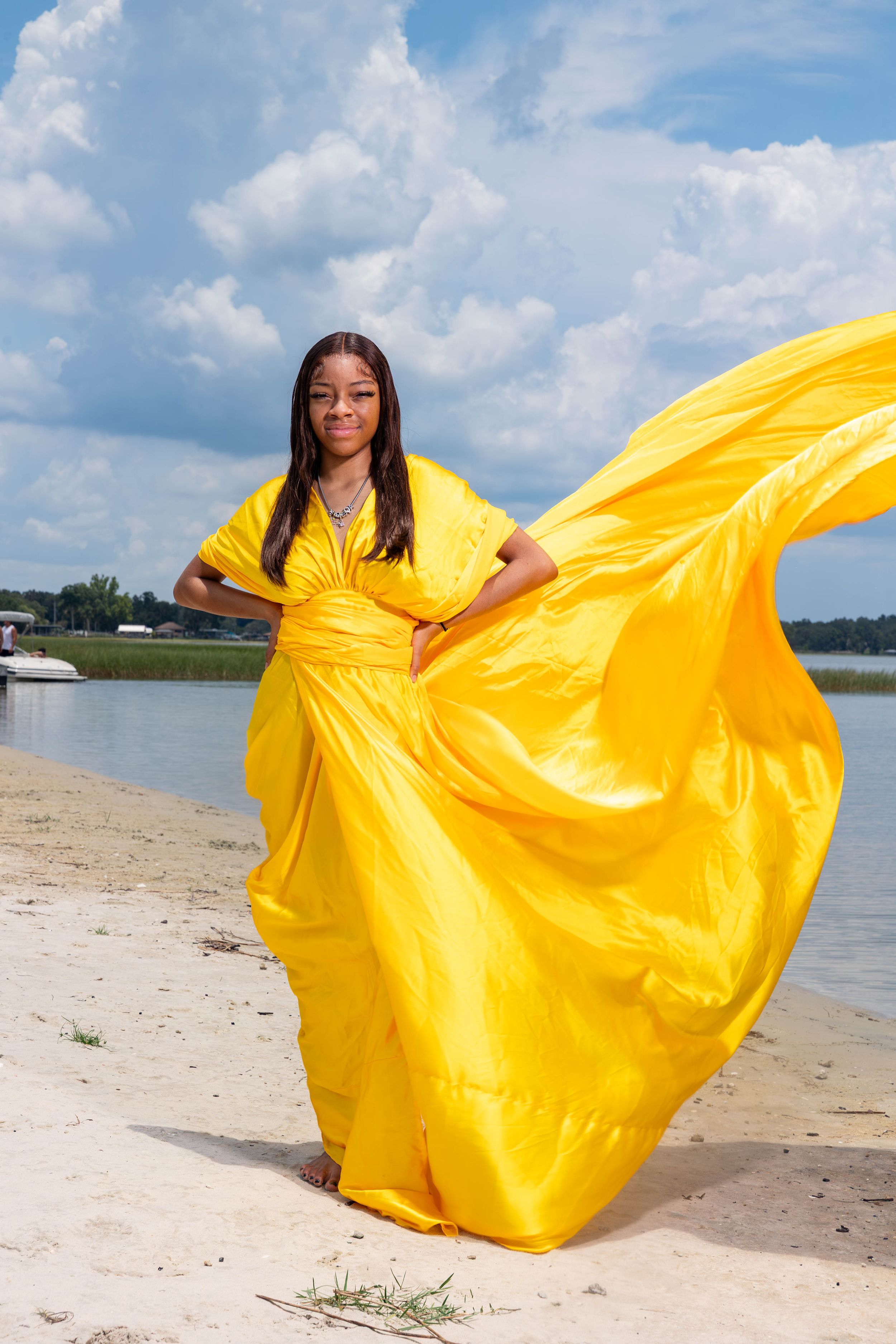 A Black woman in a flowing yellow gown poses confidently on a sandy beach with the wind catching her dress.