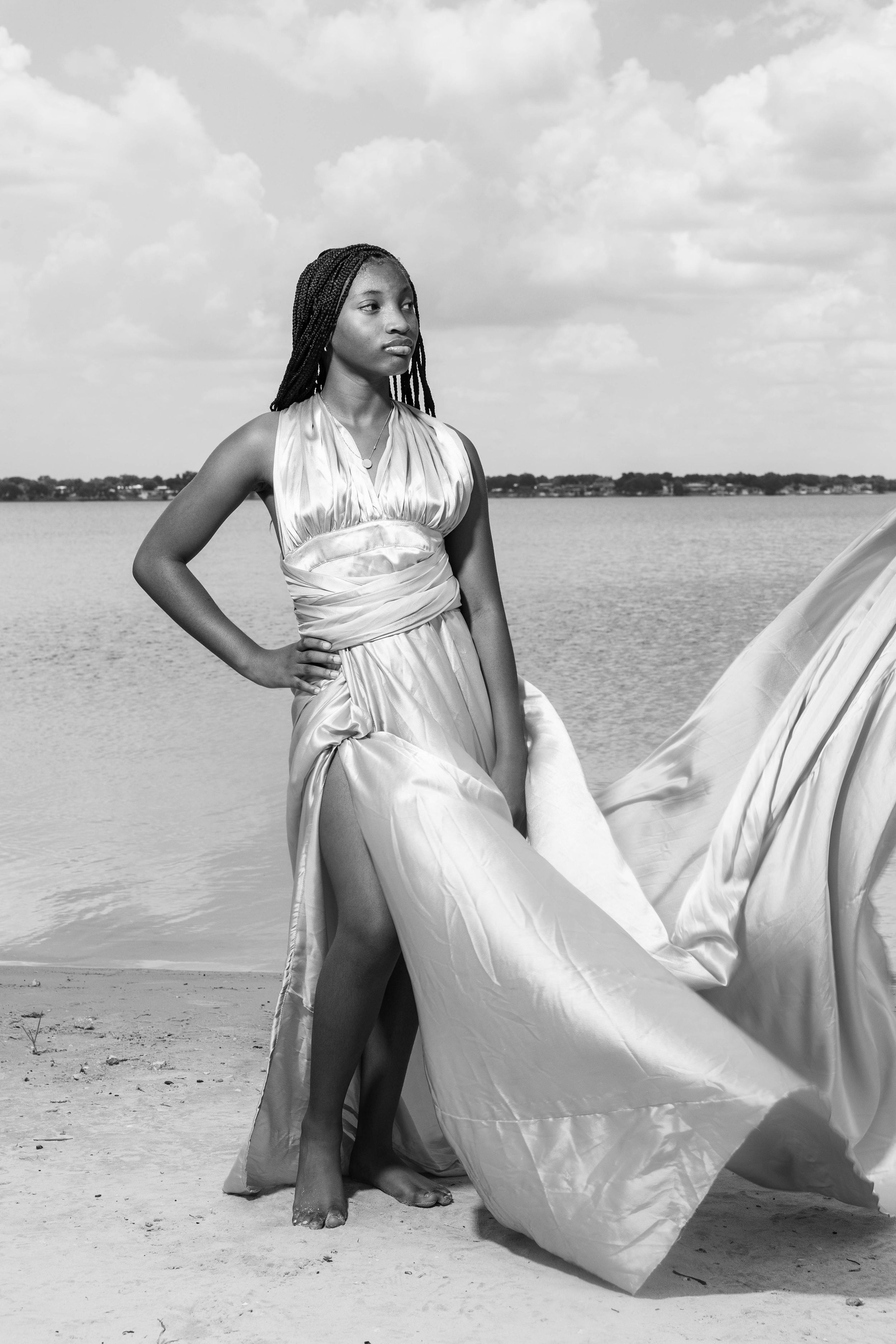 Black and white photo of a young woman in a flowing satin gown posing gracefully on a sandy beach.