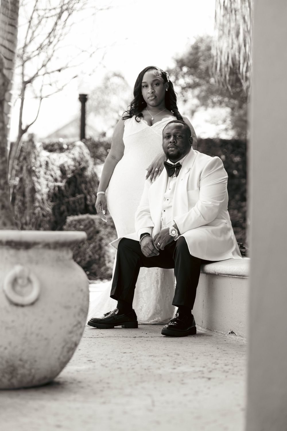 Black and white photo of a couple in wedding attire, woman in white gown standing behind seated man in white tuxedo.