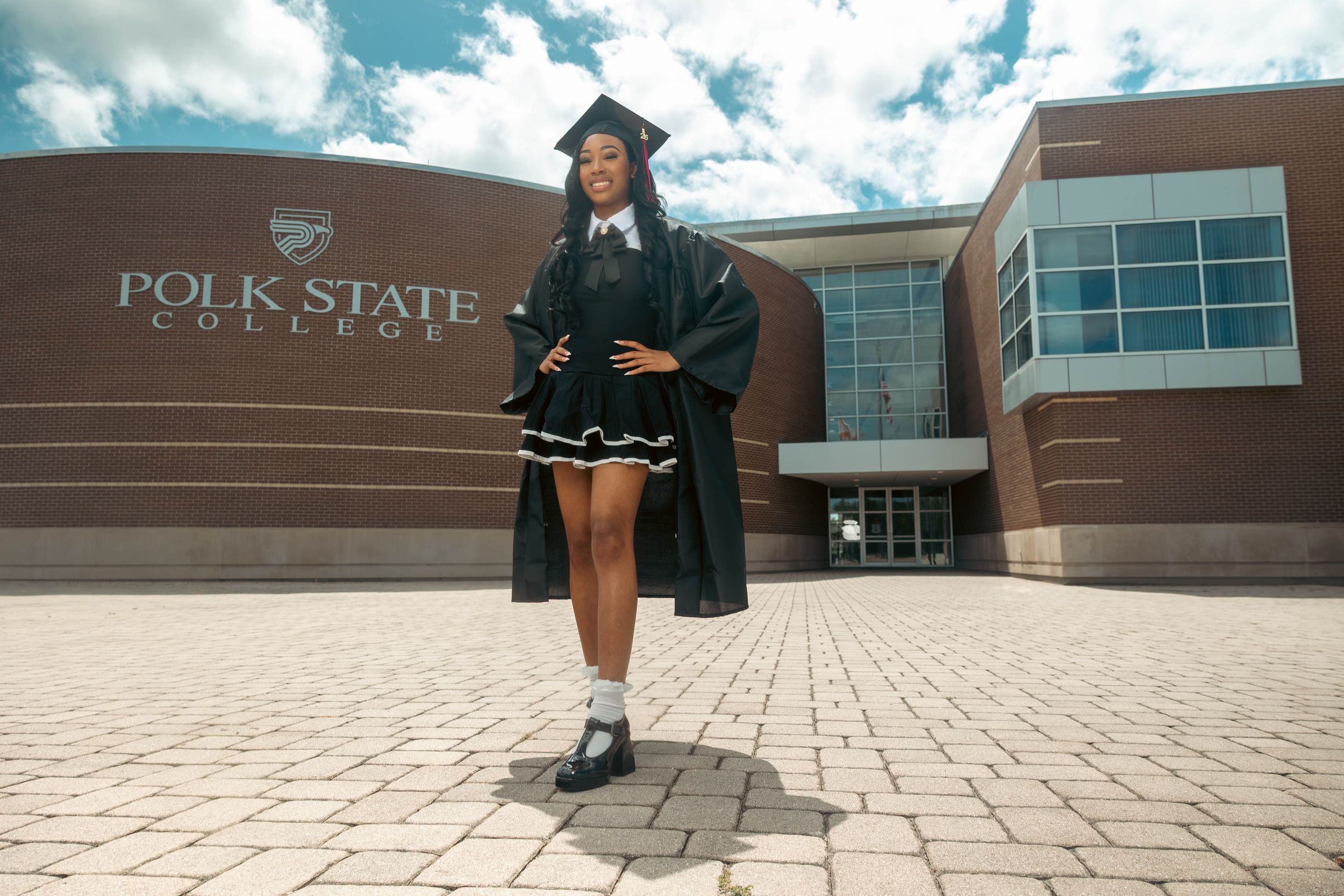 Graduate in cap and gown posing proudly in front of Polk State College building on a sunny day.