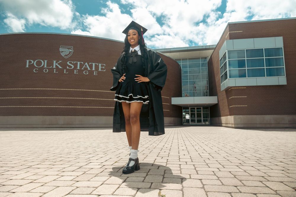 Graduate in cap and gown posing proudly in front of Polk State College building on a sunny day.