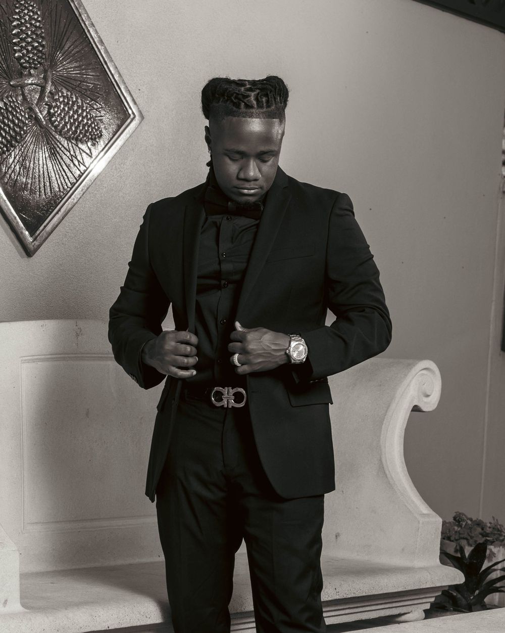 A stylish man in a black suit adjusts his Ferragamo belt, wearing a watch, with braided hair, in a black and white photo.