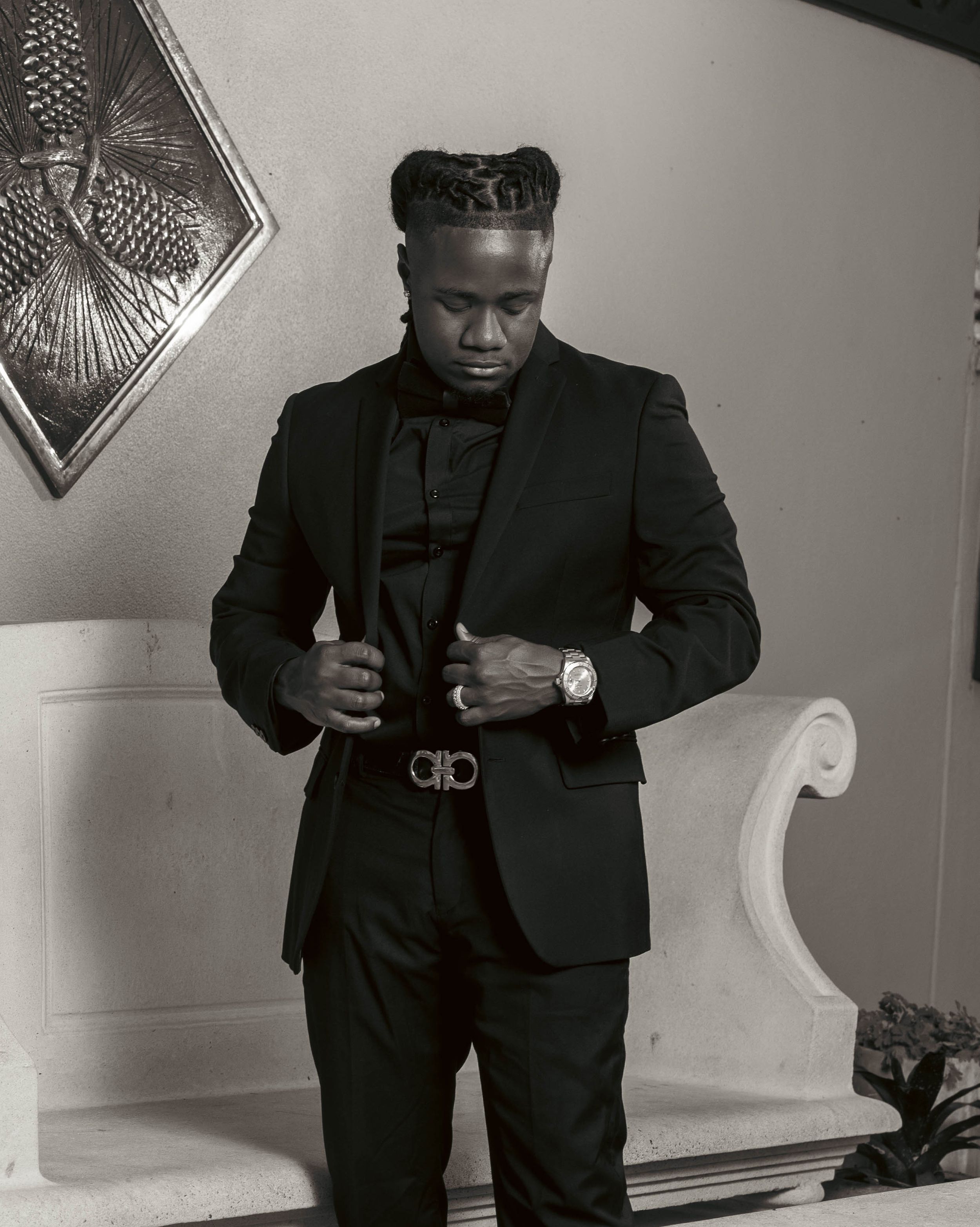 A stylish man in a black suit adjusts his Ferragamo belt, wearing a watch, with braided hair, in a black and white photo.
