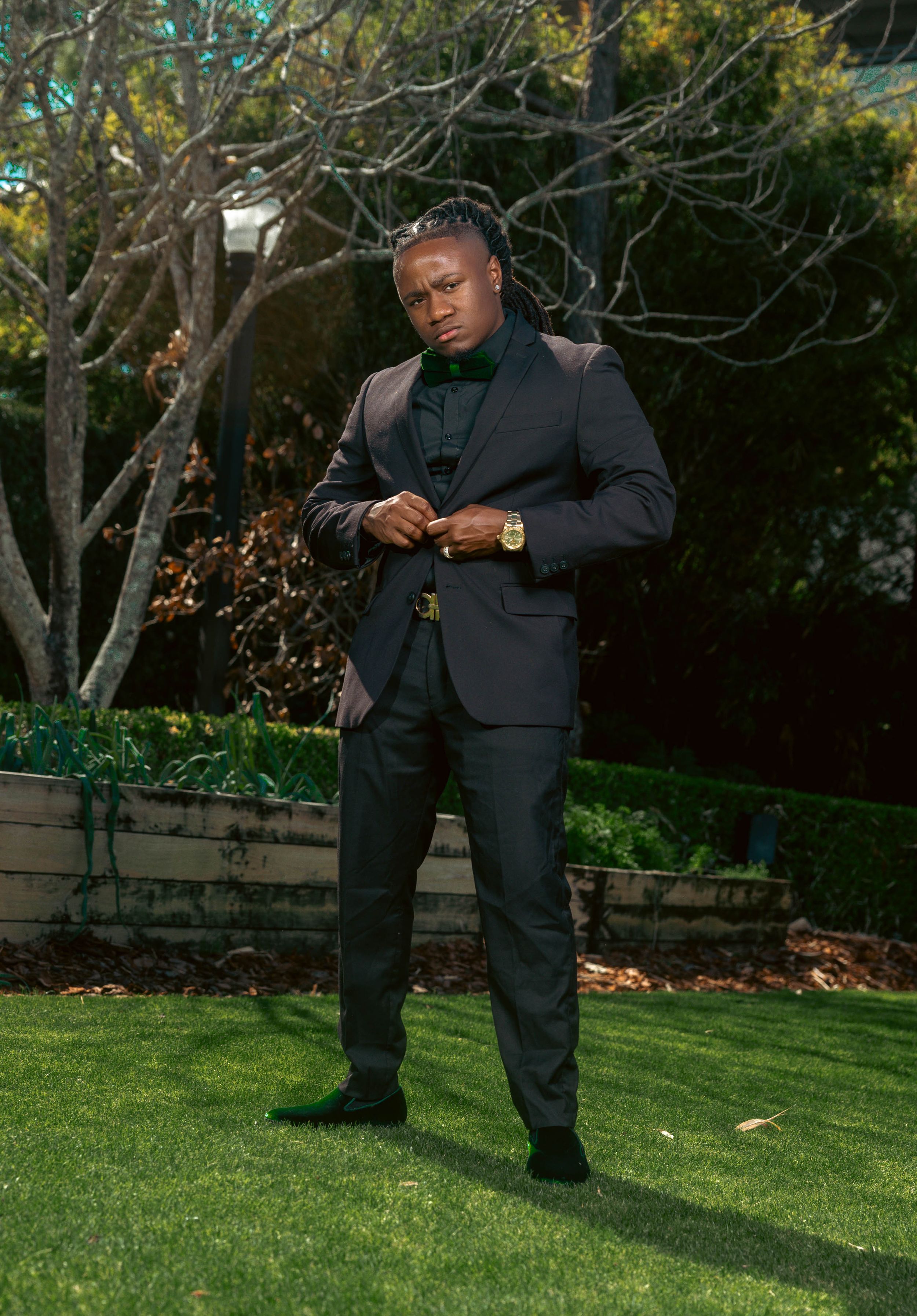 Man in dark suit posing confidently outdoors on green lawn with trees and garden beds in background.