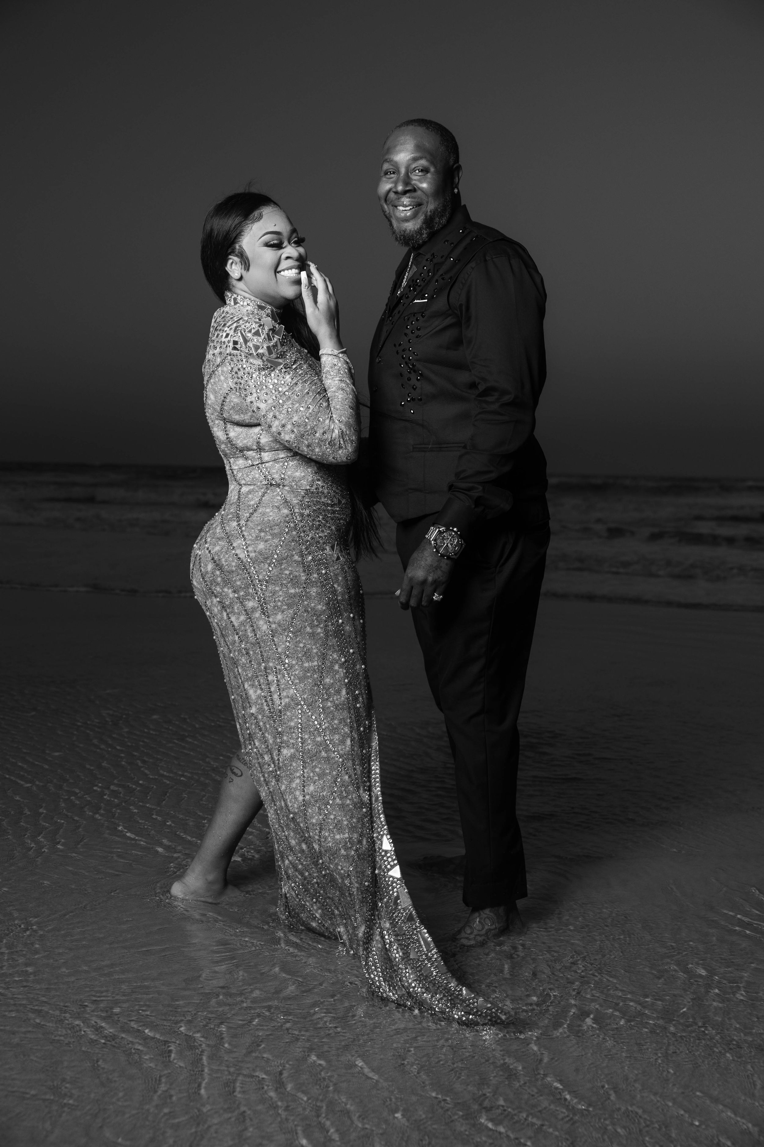 Black and white photo of a couple posing on a beach at dusk, woman in sequin gown, man in black suit.
