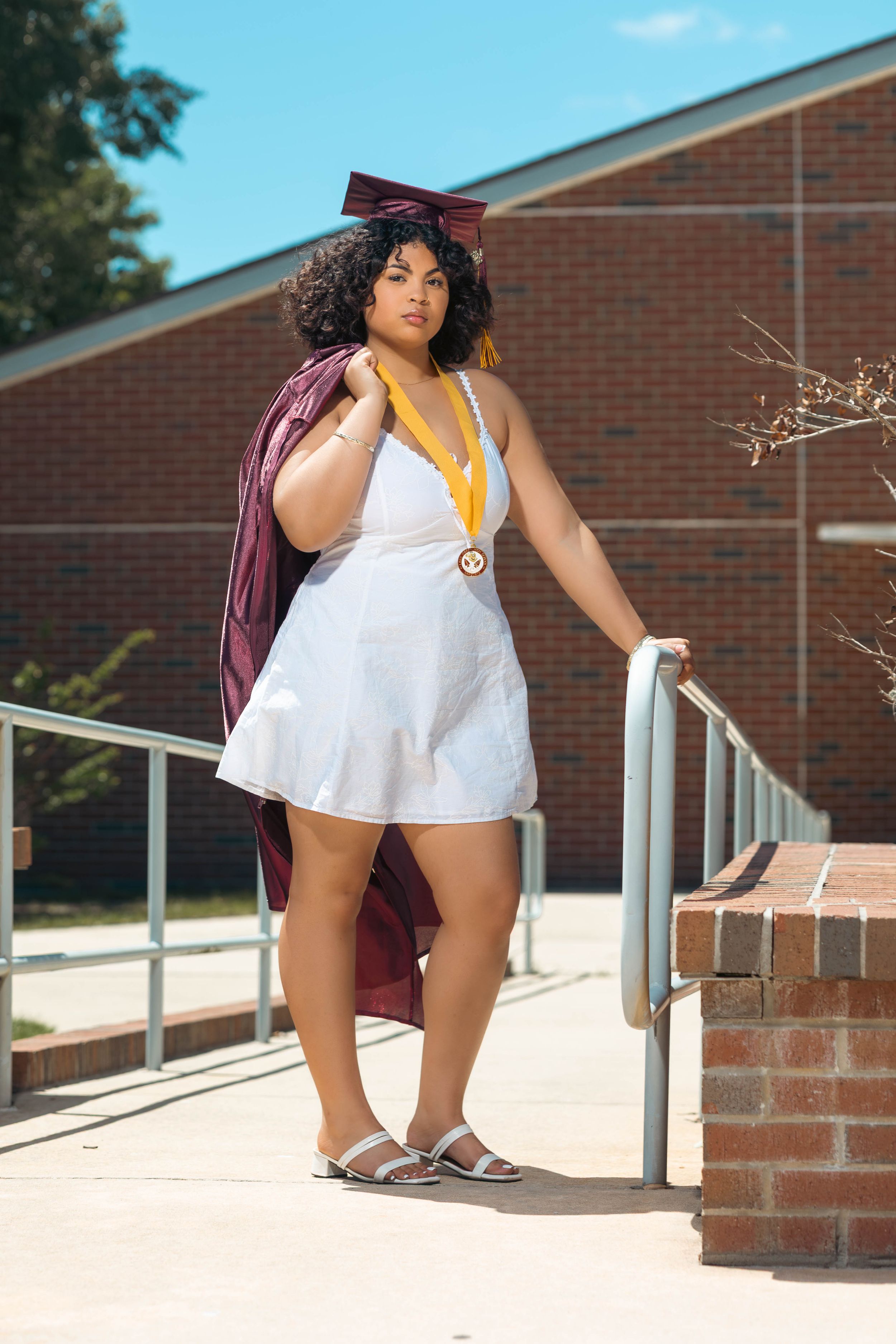 Young woman in white dress with maroon graduation cap and gown, wearing gold medal, posing by metal railings outside brick building.