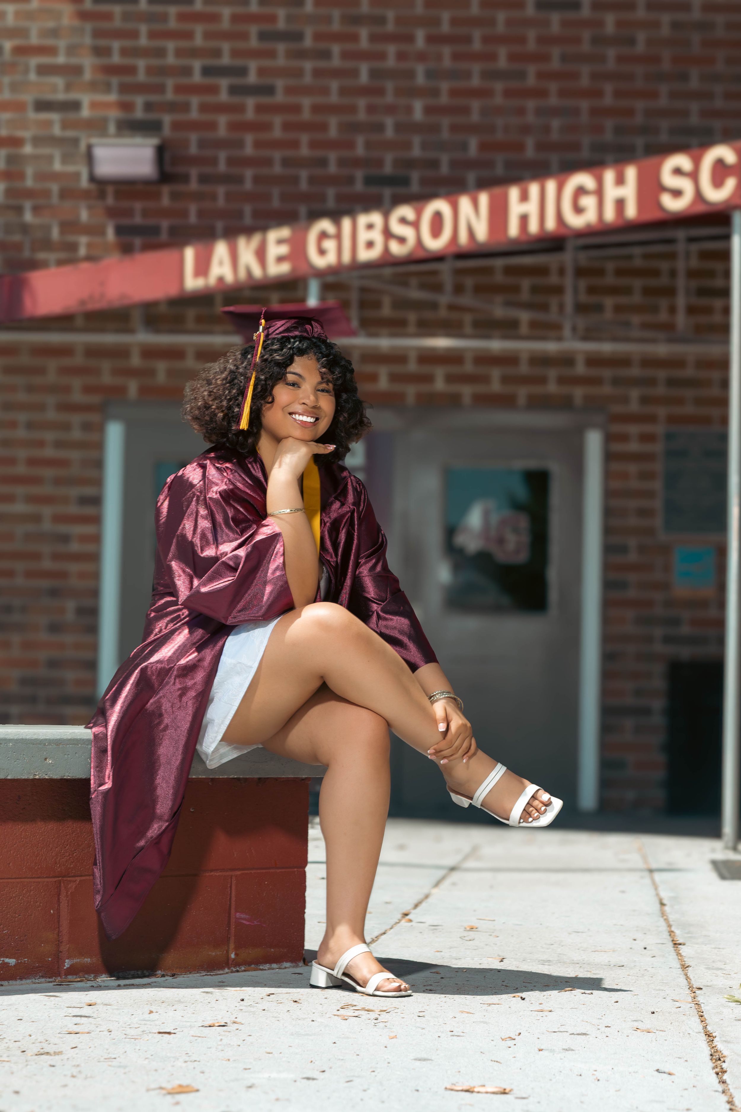 A smiling graduate in maroon cap and gown sits outside Lake Gibson High School on a sunny day.