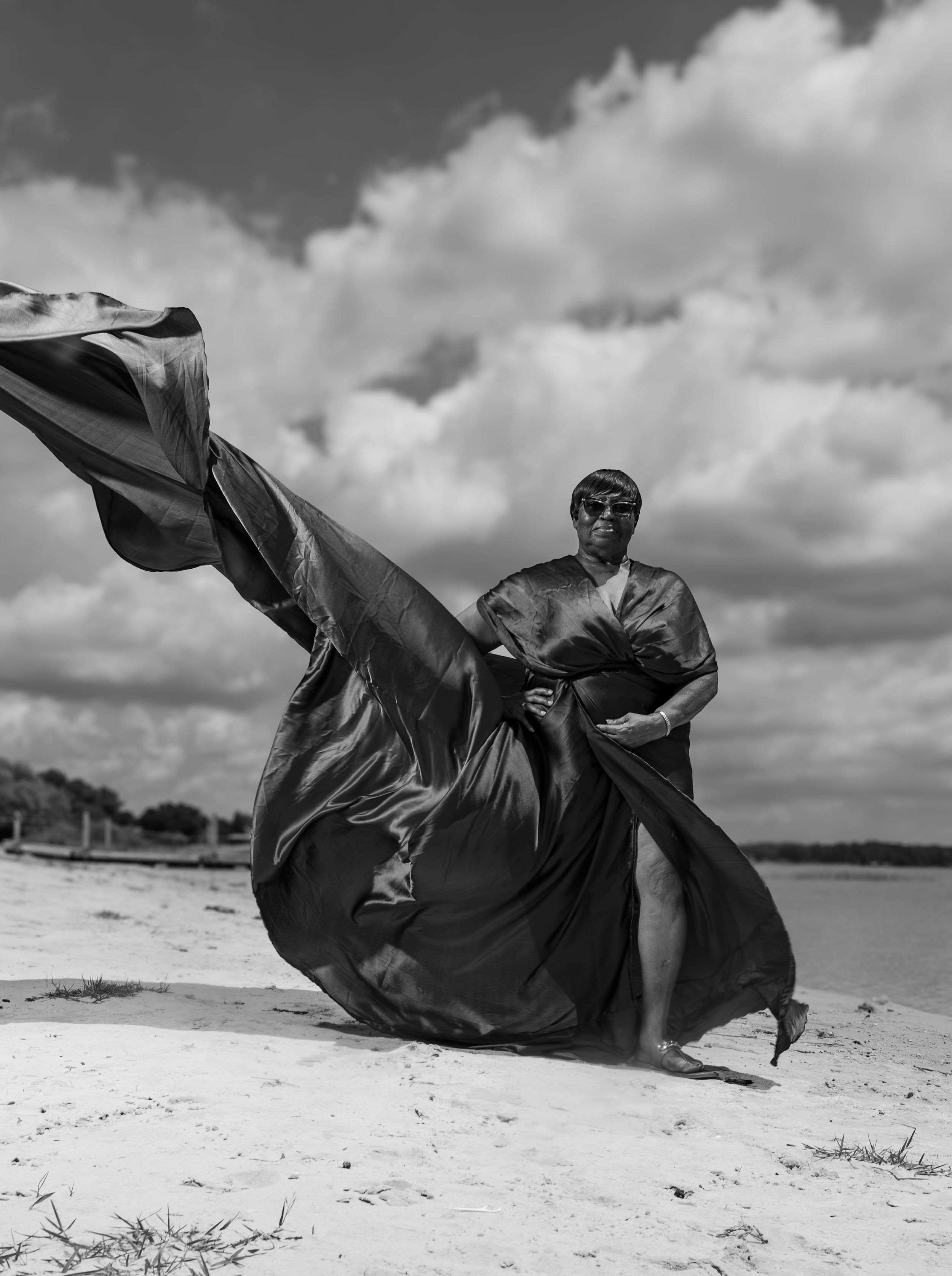 Black and white photo of a person in flowing fabric on a beach, dramatic clouds behind them.