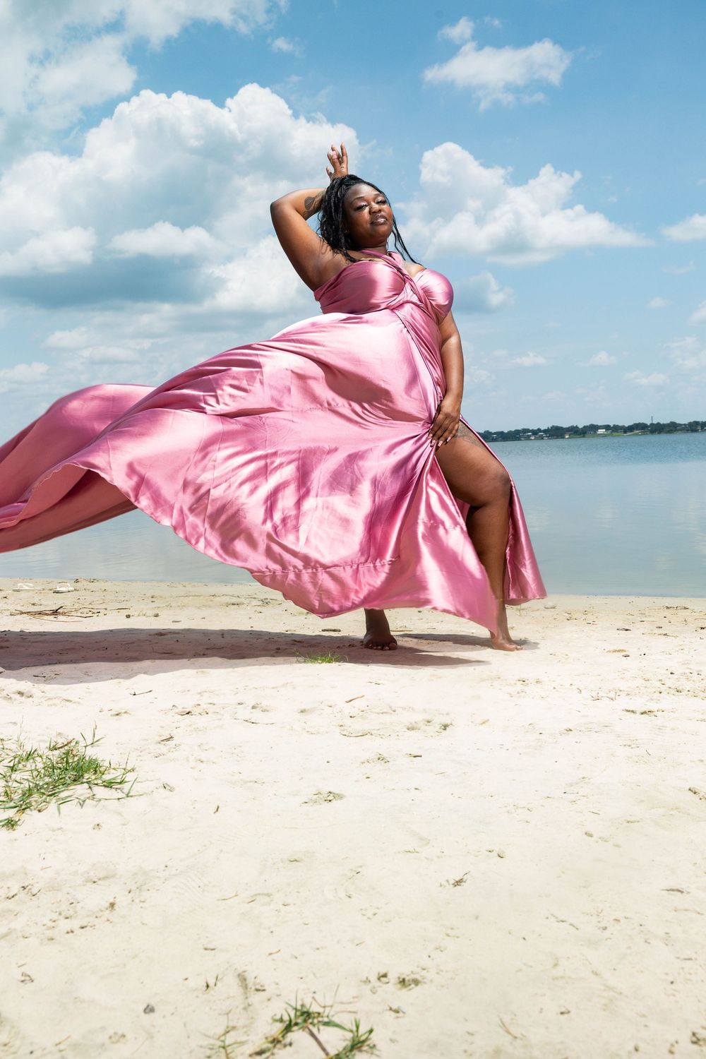 A plus-size woman in a flowing pink satin gown poses gracefully on a sandy beach under a blue sky.