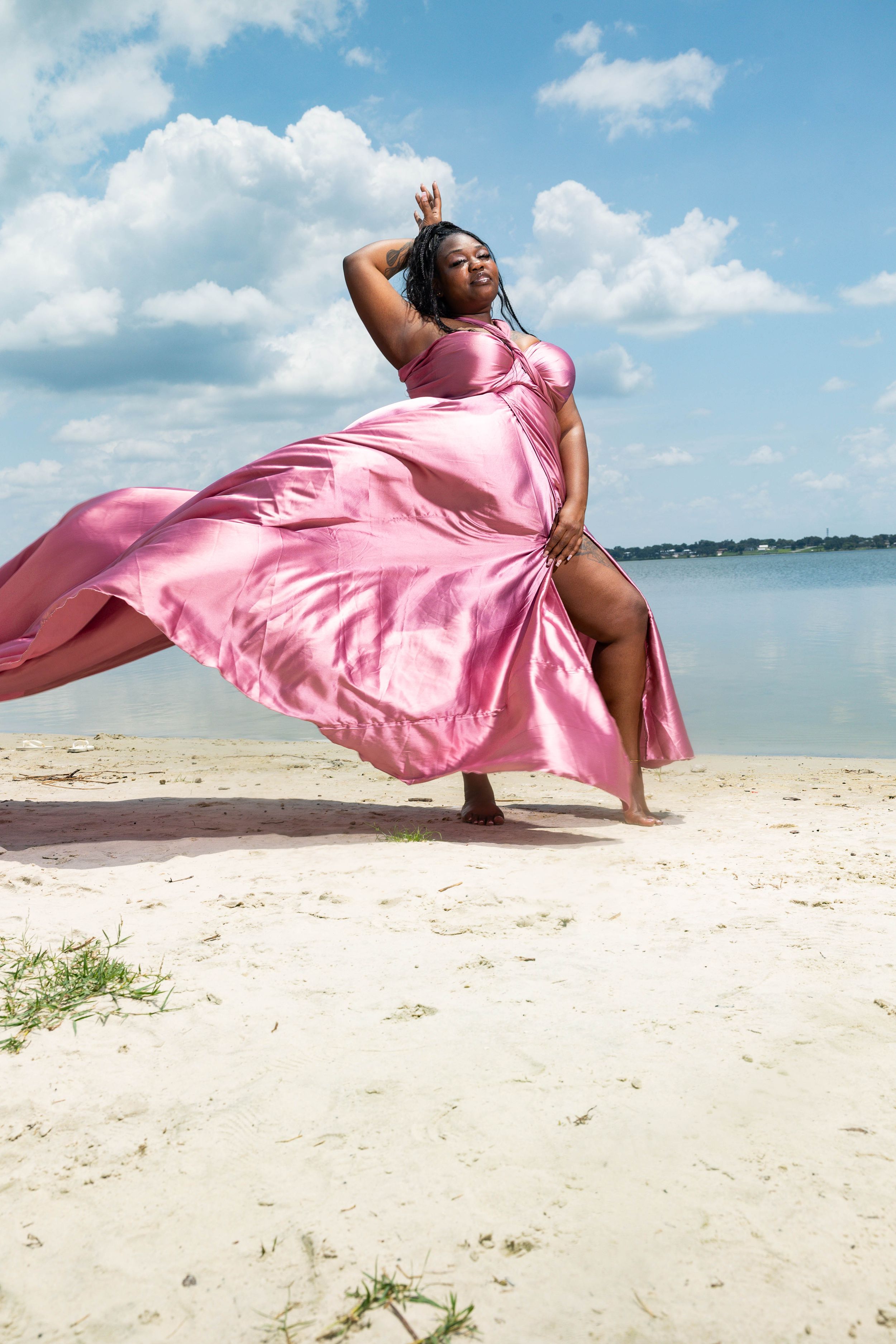 A plus-size woman in a flowing pink satin gown poses gracefully on a sandy beach under a blue sky.