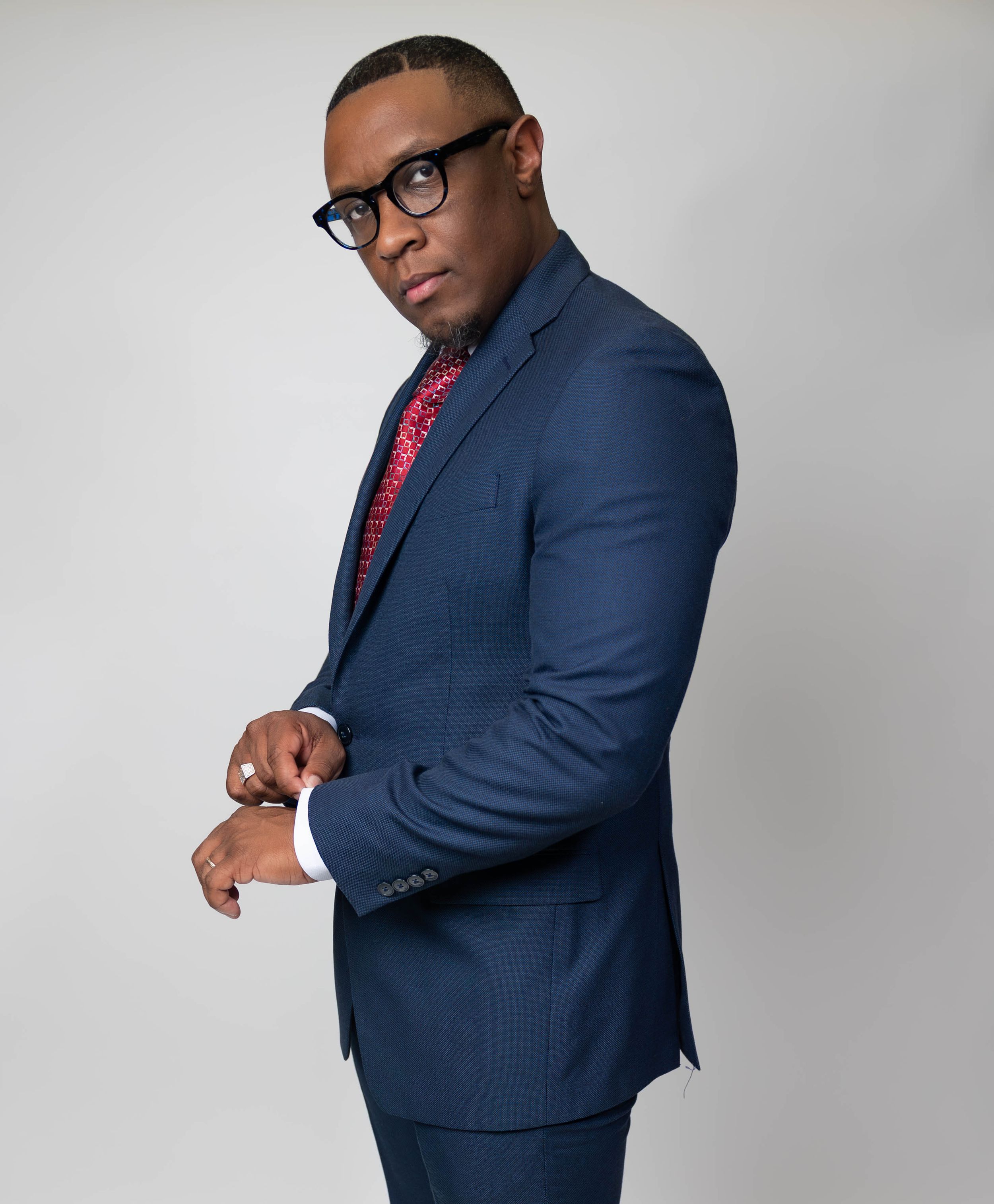 Man in navy blue suit with red patterned tie and black glasses poses confidently against gray background.