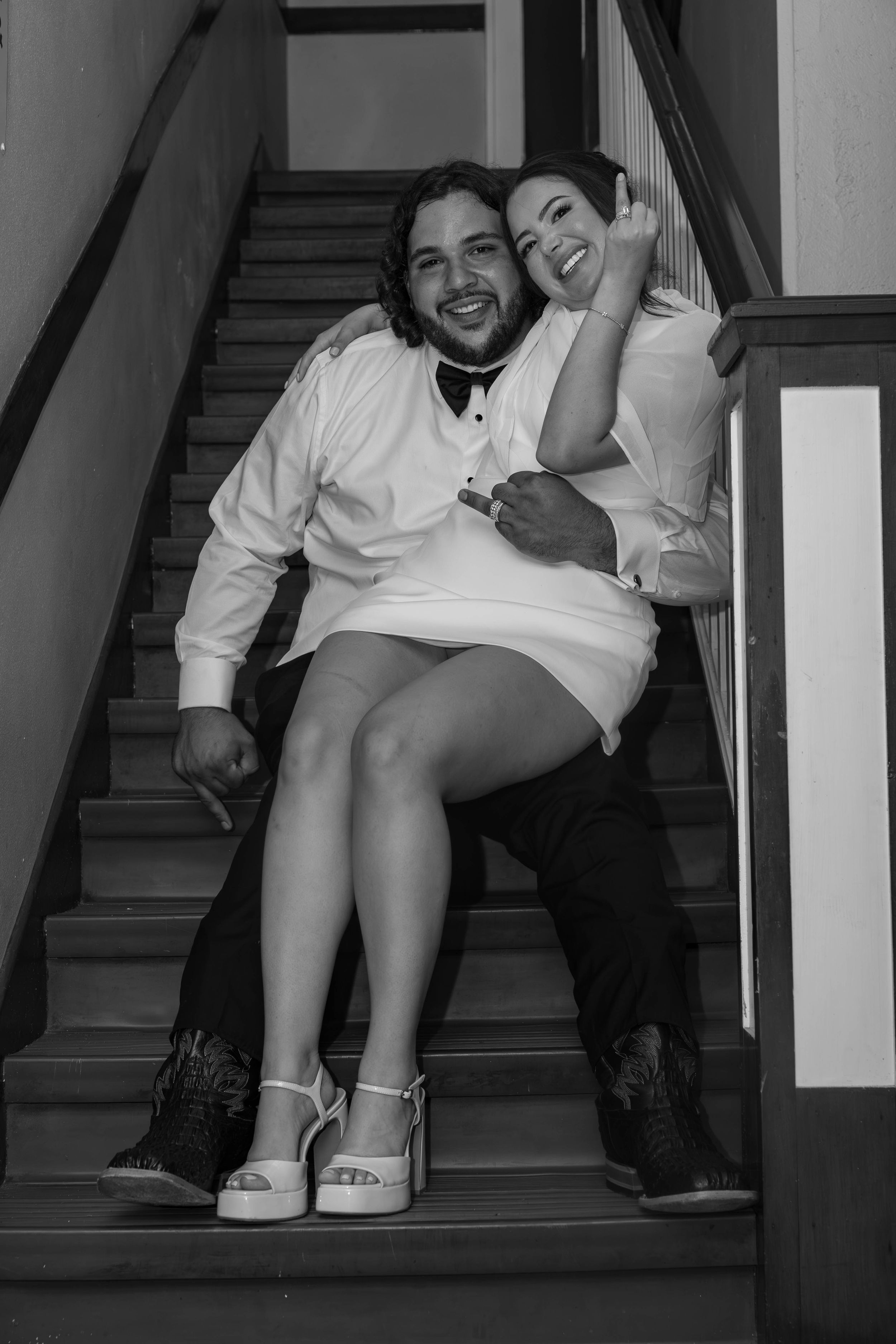 Black and white photo of a smiling couple sitting on stairs, dressed in white formal attire, sharing a joyful embrace.
