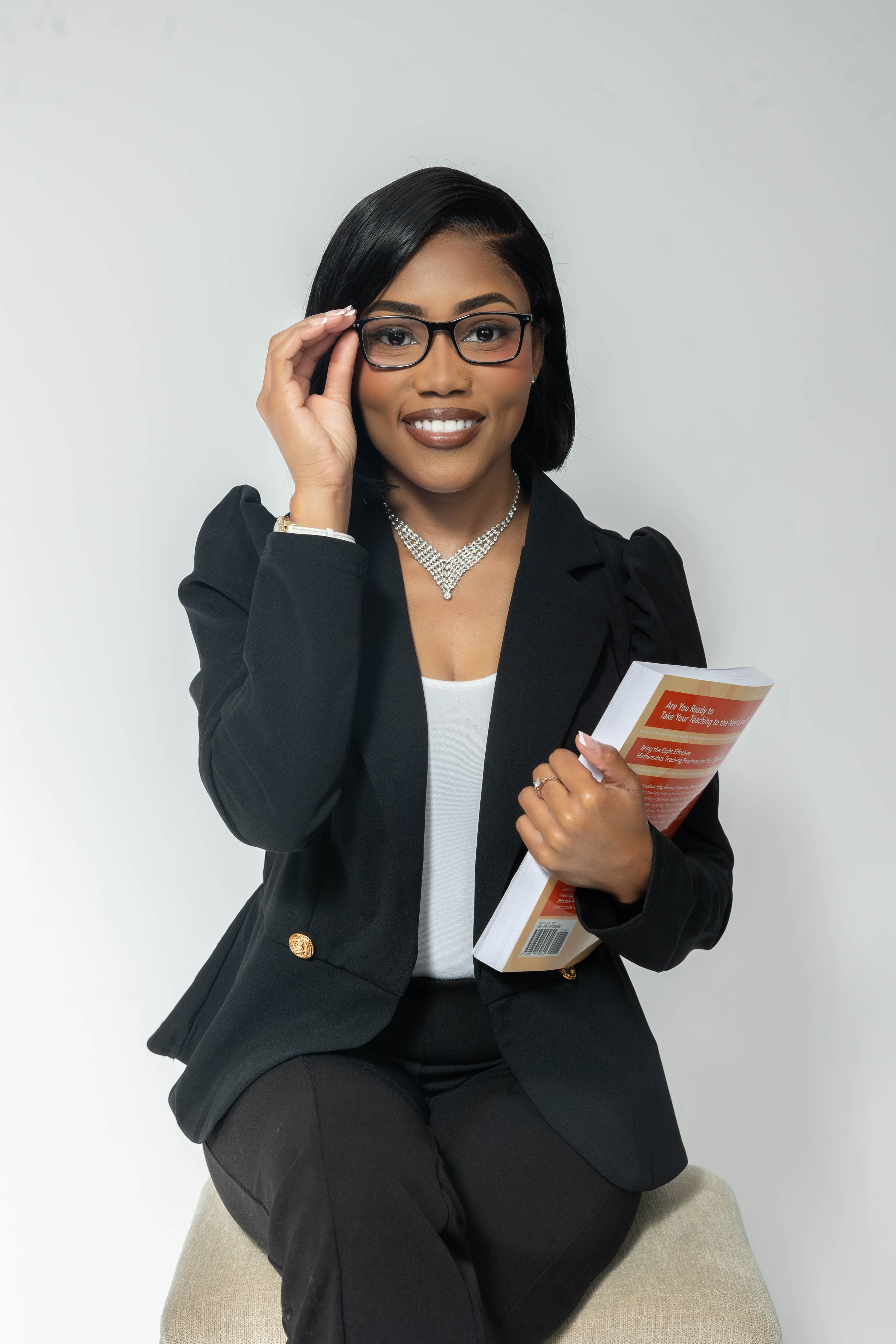 Smiling professional Black woman in black blazer and glasses holding a book, seated against white background.