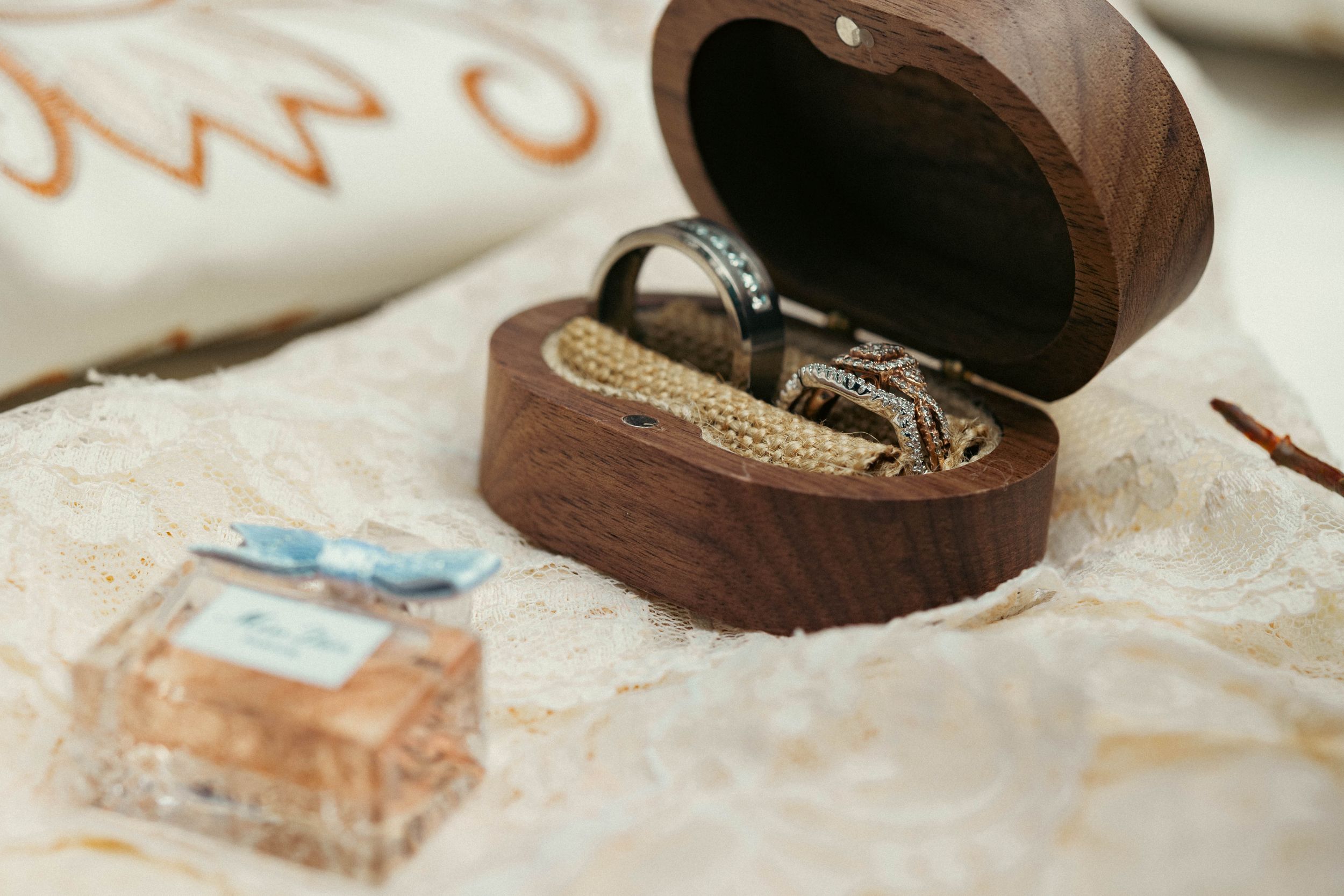 Wooden ring box with wedding bands and gold chain beside a Dior perfume bottle on lace fabric.