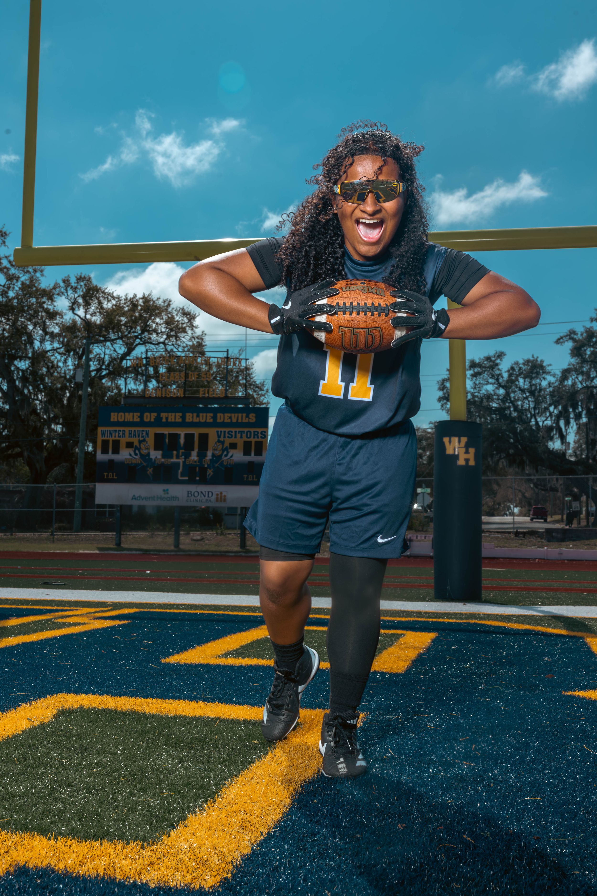 Female football player #11 in navy uniform holds ball on field, smiling confidently in front of goal post on sunny day.