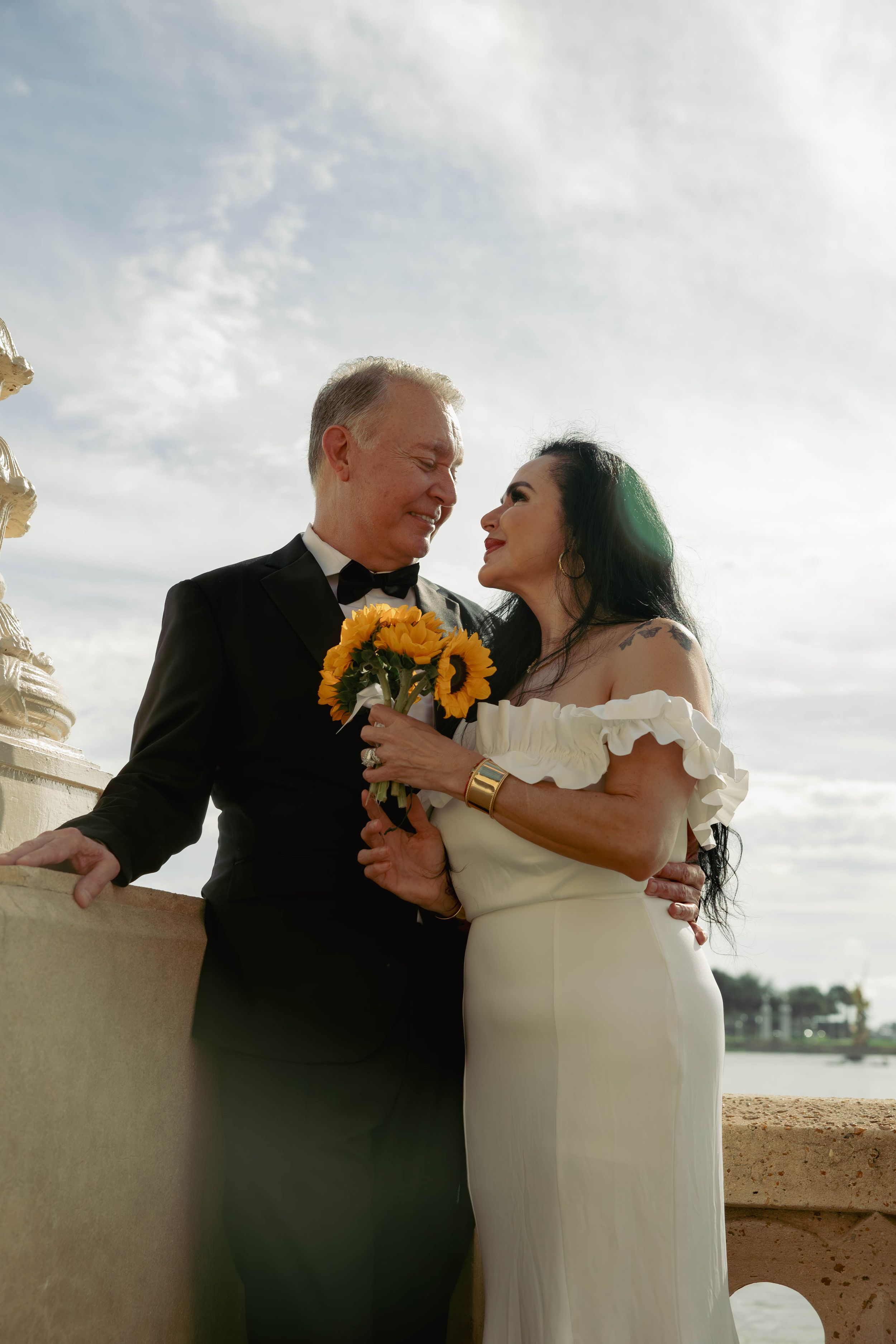 A bride and groom share a romantic moment on a balcony, she holding sunflowers in a white gown, he in a black tuxedo.