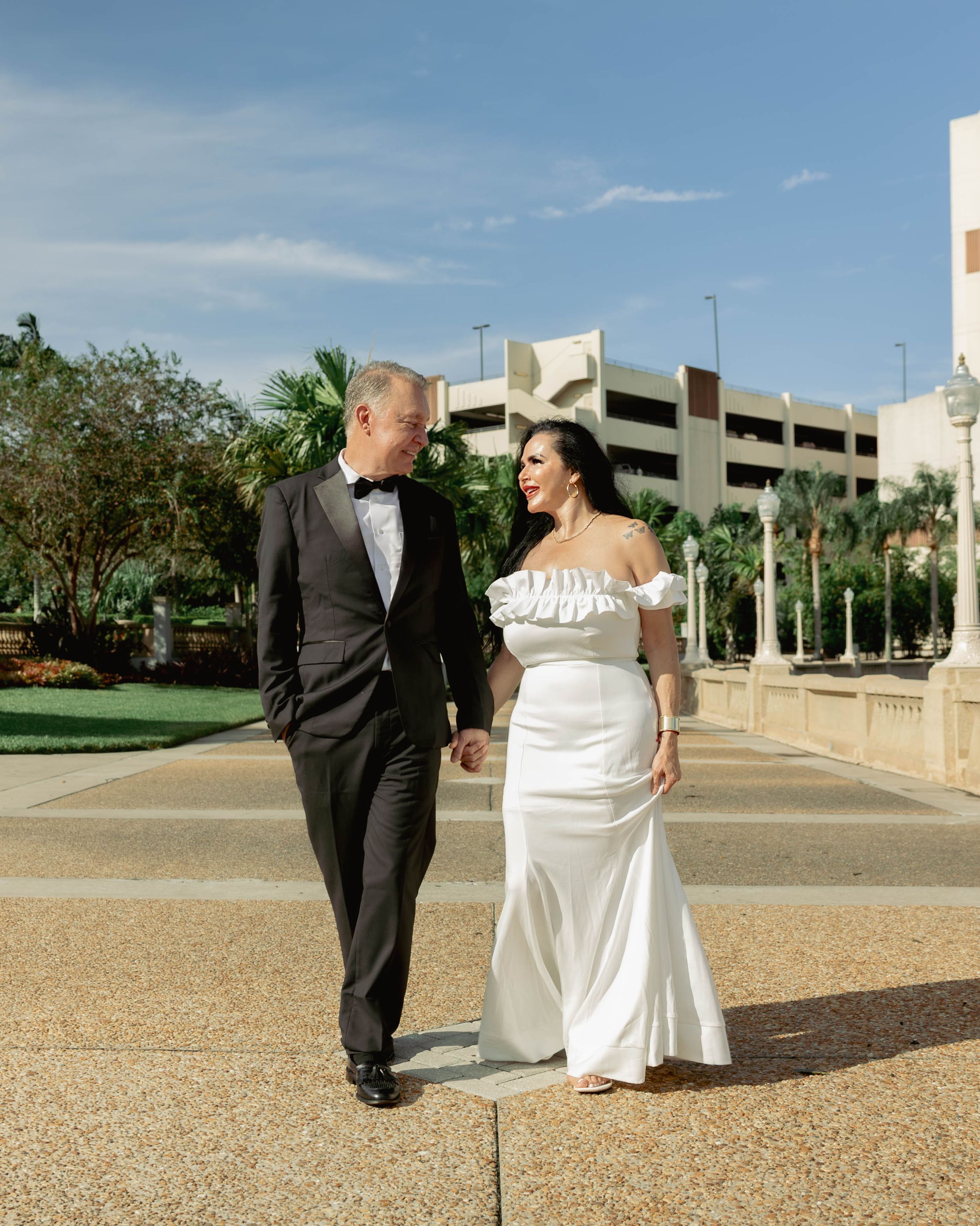 Couple in wedding attire walking hand in hand outdoors, groom in tuxedo and bride in white off-shoulder gown.