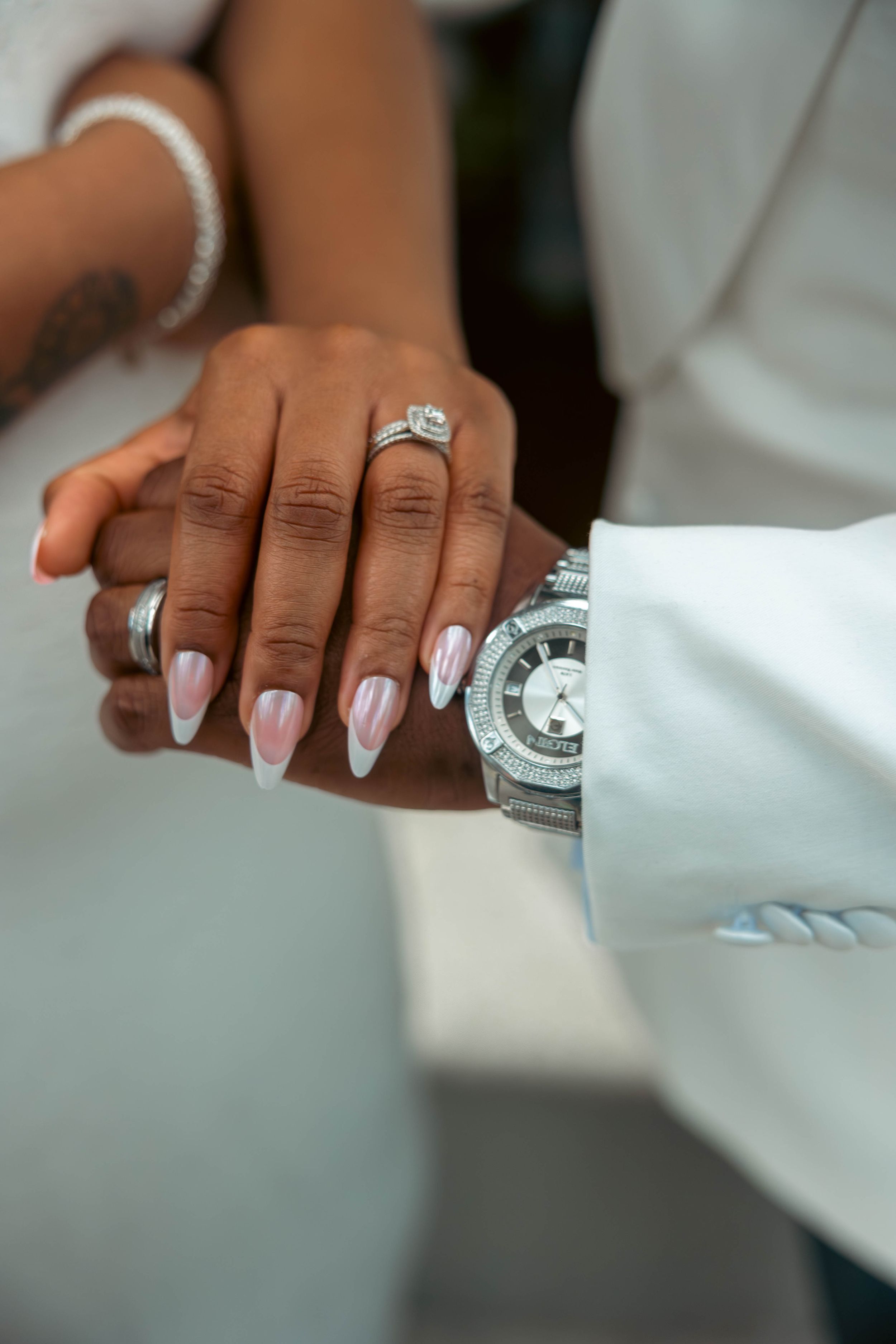 Close-up of a couple's hands showing a diamond engagement ring, French tip nails, and a silver luxury watch on wedding day.