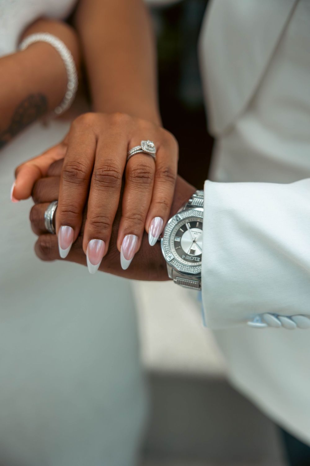 Close-up of a couple's hands showing a diamond engagement ring, French tip nails, and a silver luxury watch on wedding day.